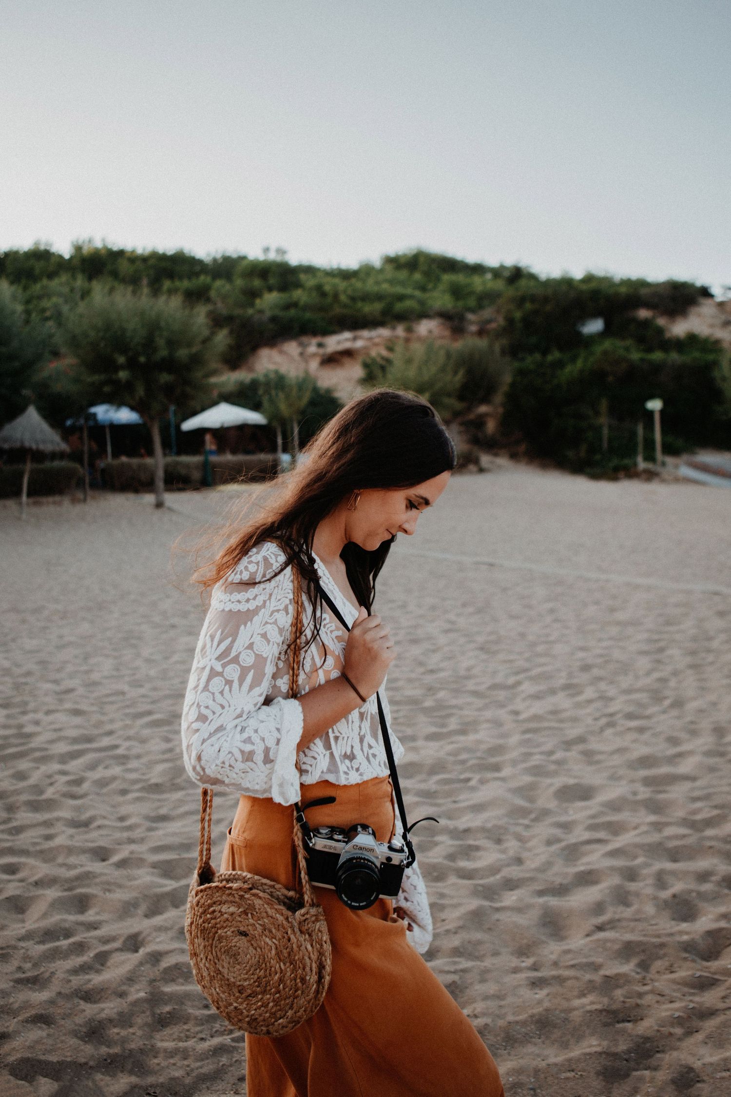 Bohemian woman in white lace top and orange pants walks on sandy beach carrying vintage camera.