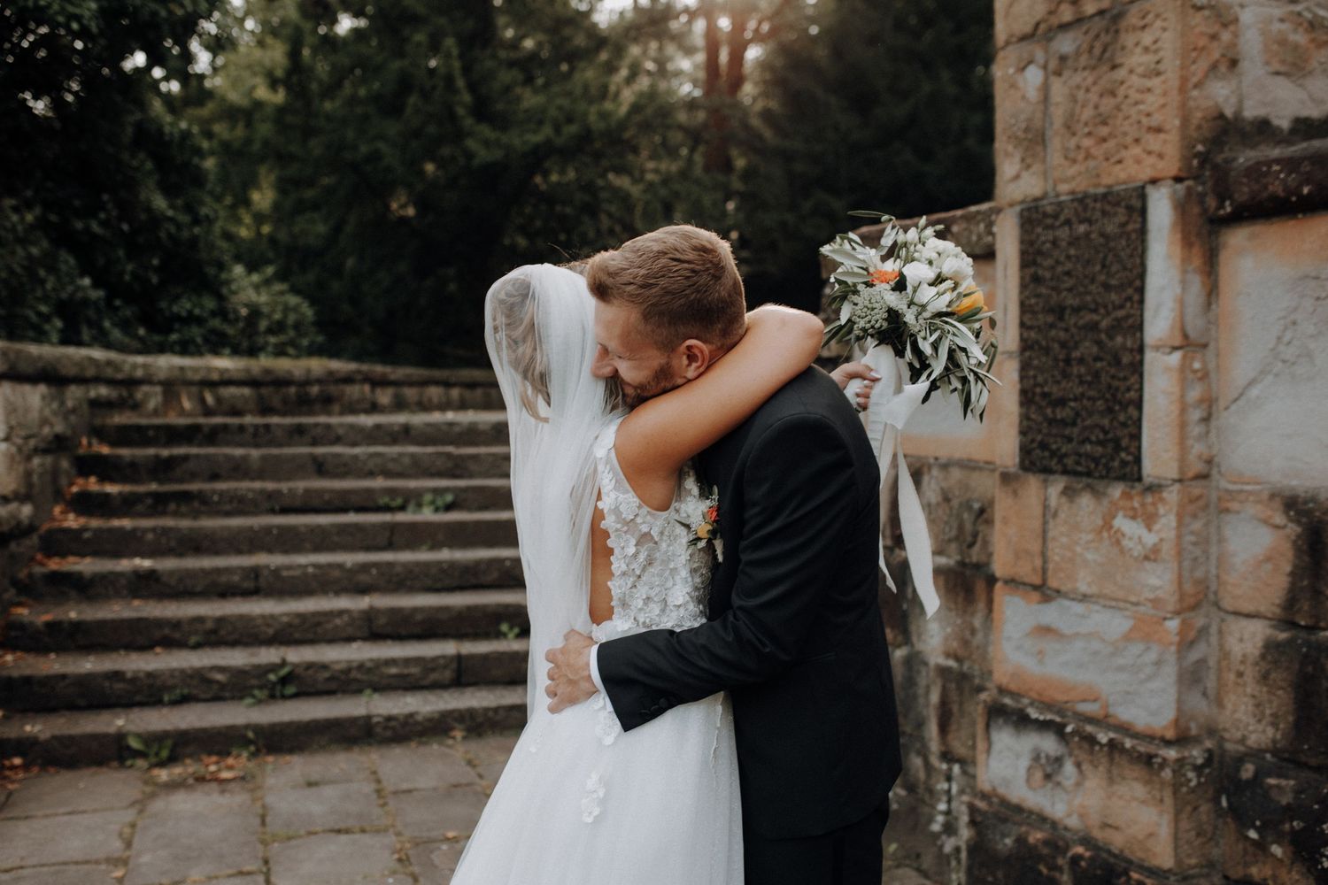 Bride and groom share a tender embrace near stone steps, bride holding a white bouquet in a lace wedding dress.
