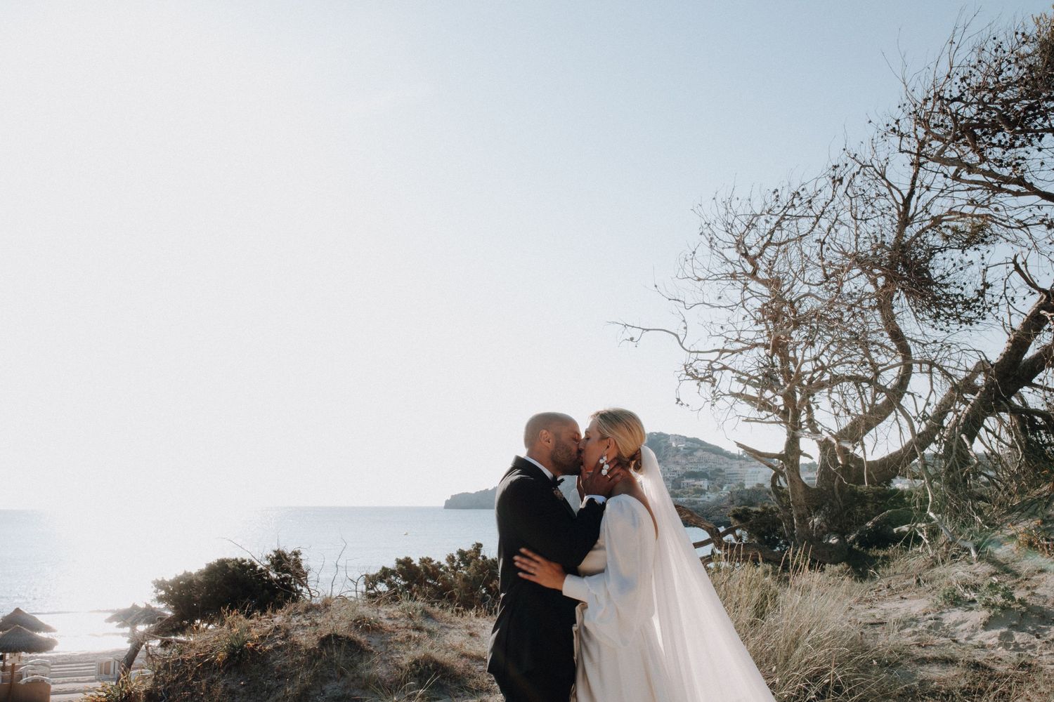 Bride and groom kissing on a scenic coastal hillside with ocean views on their wedding day.