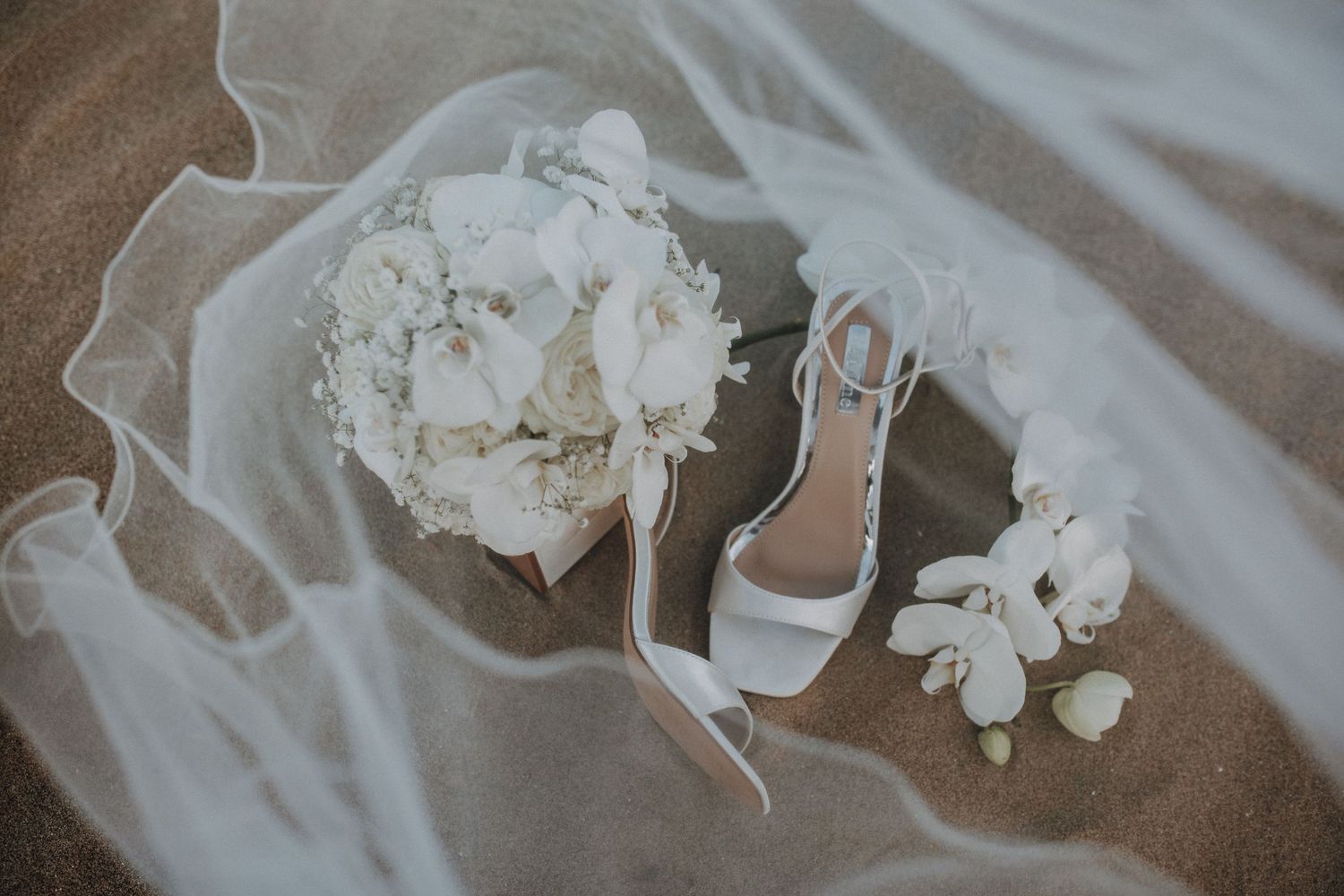 White bridal bouquet, heeled sandals, and veil arranged on sandy beach in elegant flat lay.