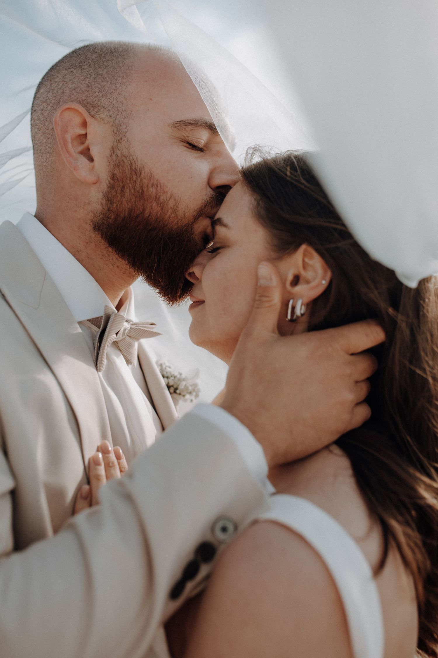 Groom in beige suit gently kisses bride on forehead under a white veil in a romantic close-up shot.