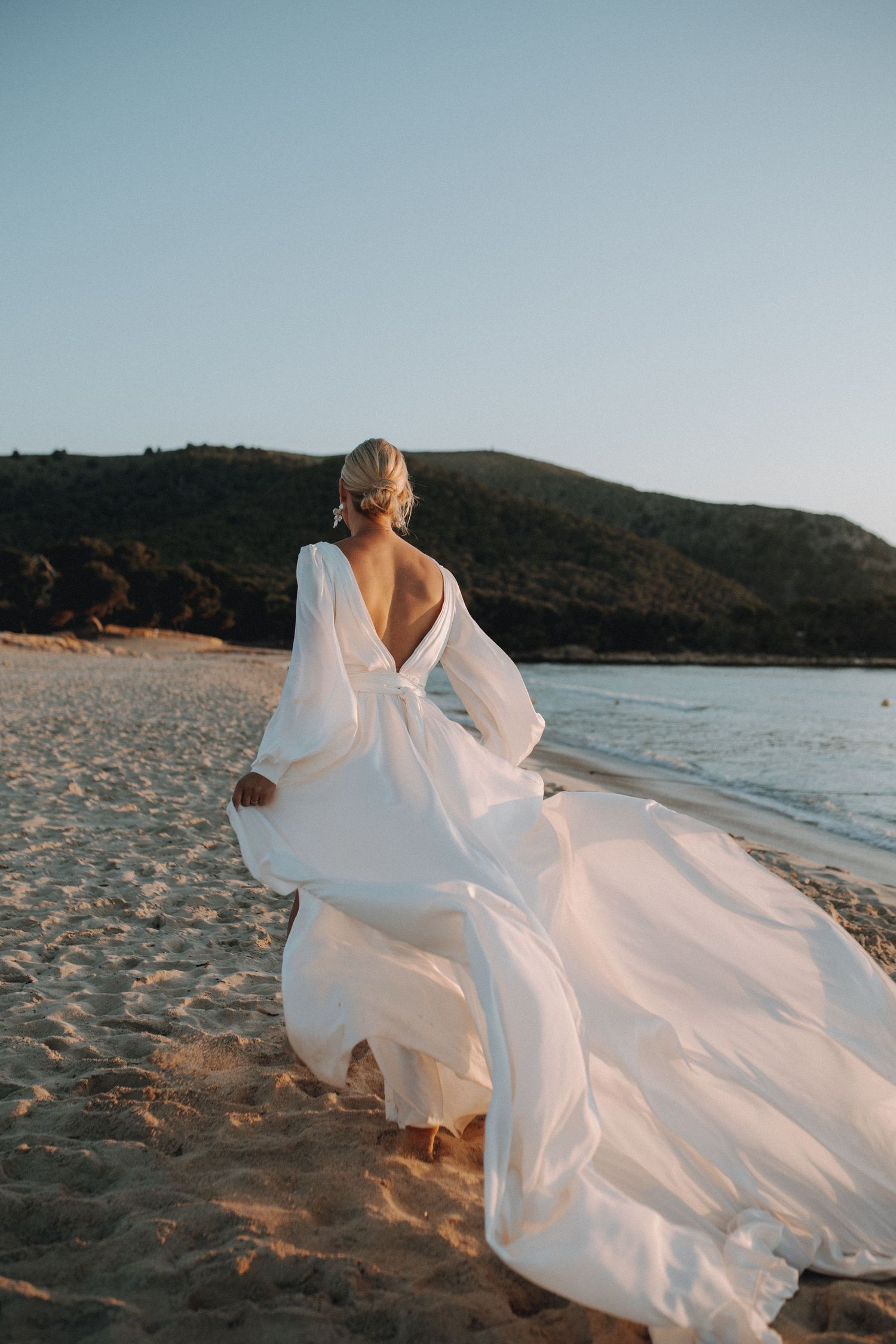 Elegant bride in flowing white backless gown walking on a sandy beach at golden hour sunset.