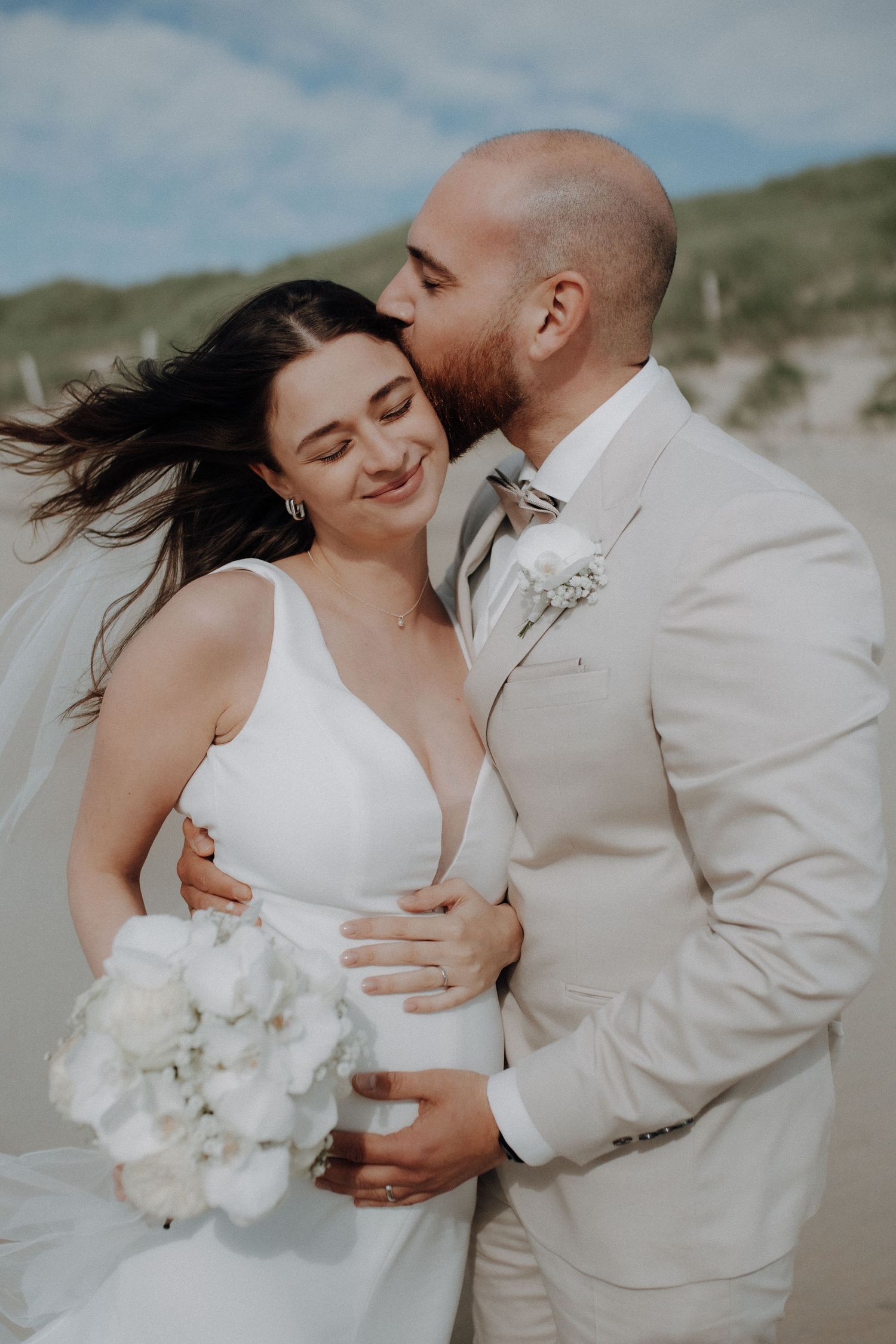 Groom in beige suit kissing bride on cheek as wind blows her hair on beach.