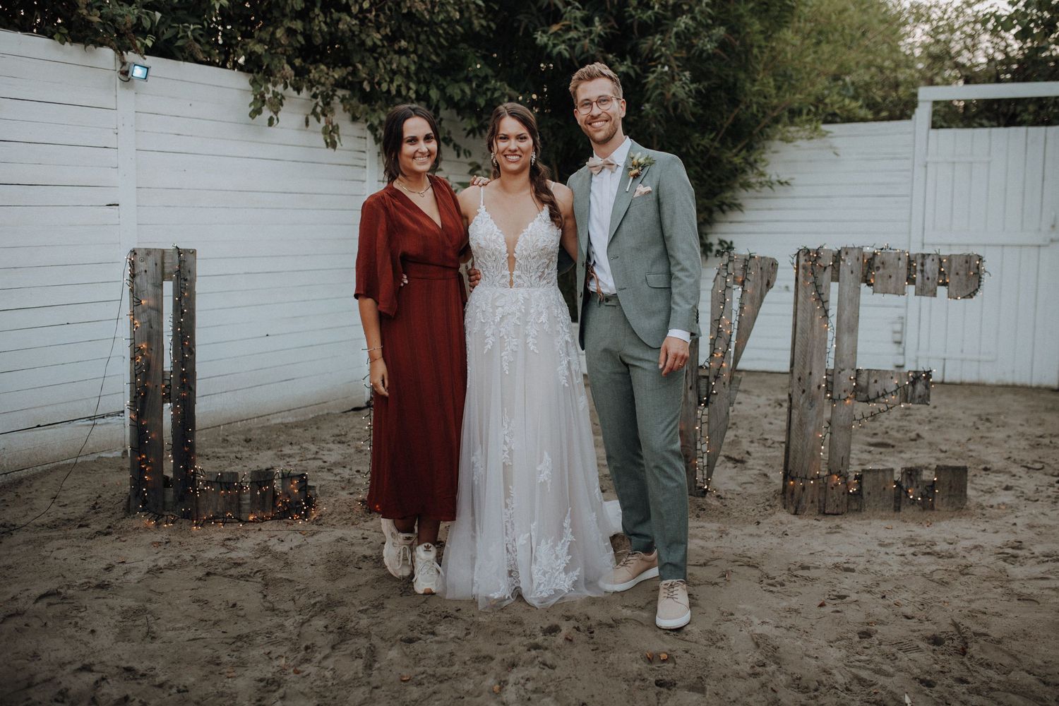 Bride and groom posing with a guest in front of decorative LOVE letters at an outdoor wedding.