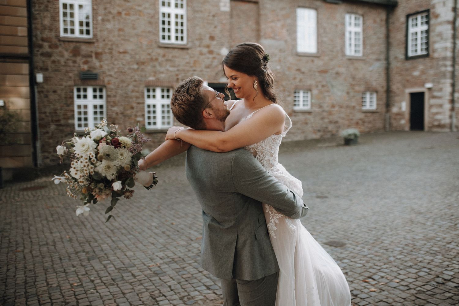 Groom lifting and spinning his bride joyfully in a cobblestone courtyard on their wedding day.
