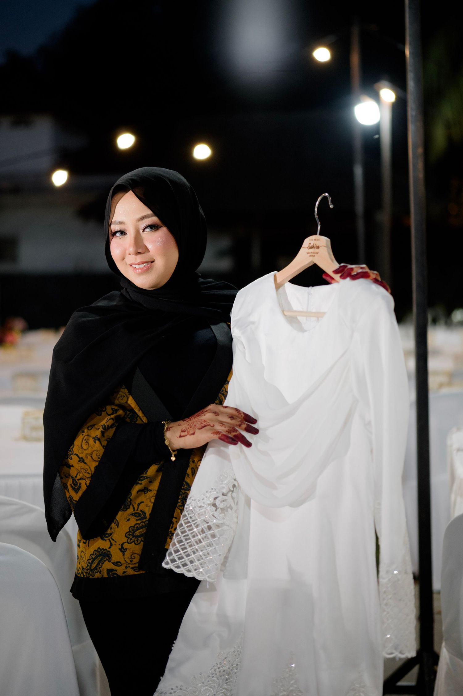 Woman in black hijab proudly displaying white wedding dress on hanger at outdoor venue.
