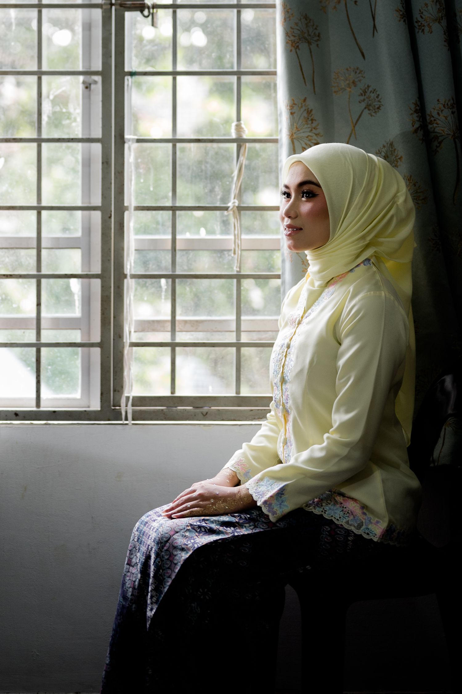 A young Muslim woman in a yellow hijab and blouse sits gracefully by a window, gazing thoughtfully outside.