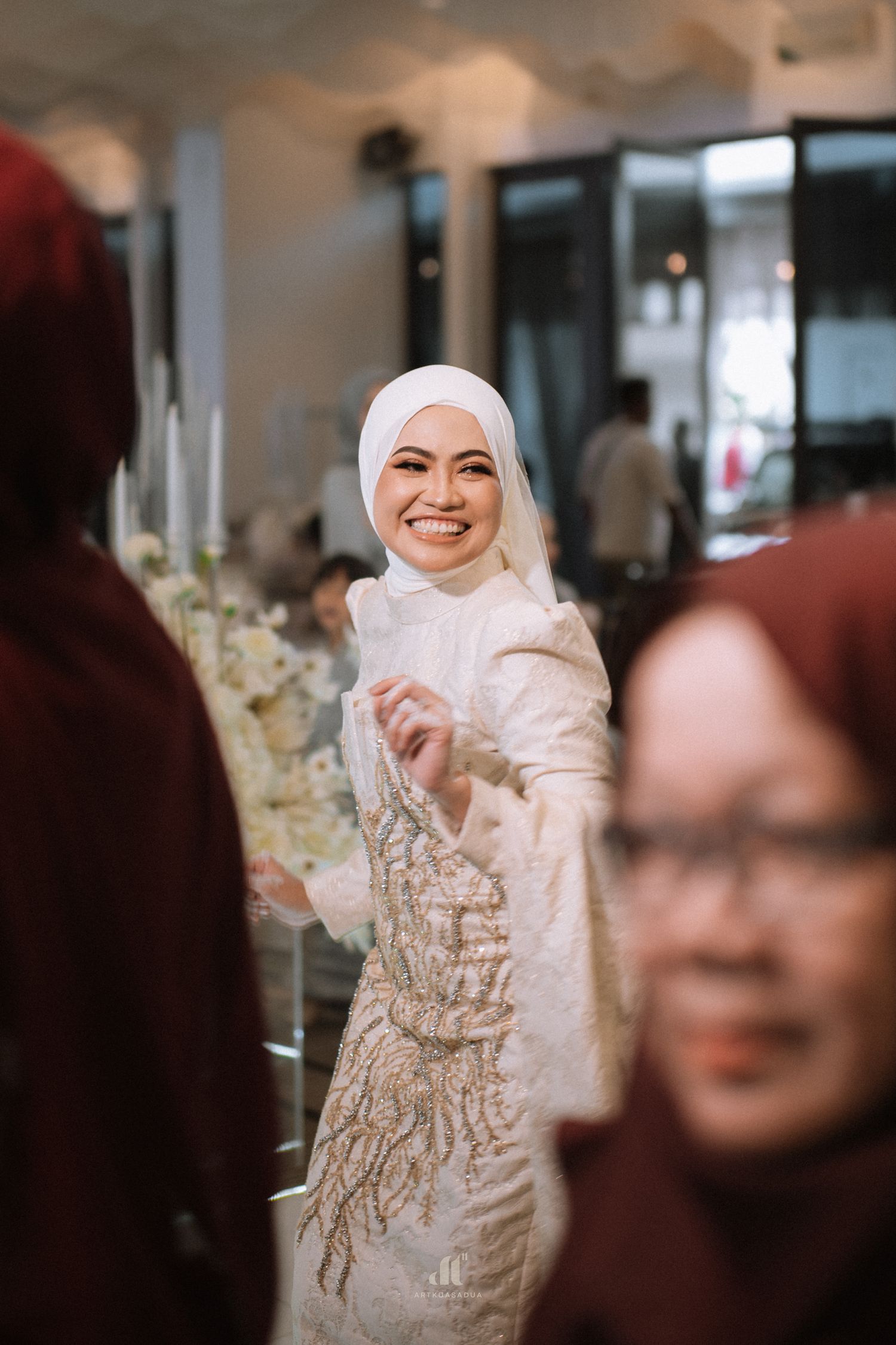 Smiling bride in white hijab and embroidered gown at a wedding reception, surrounded by guests in maroon hijabs.