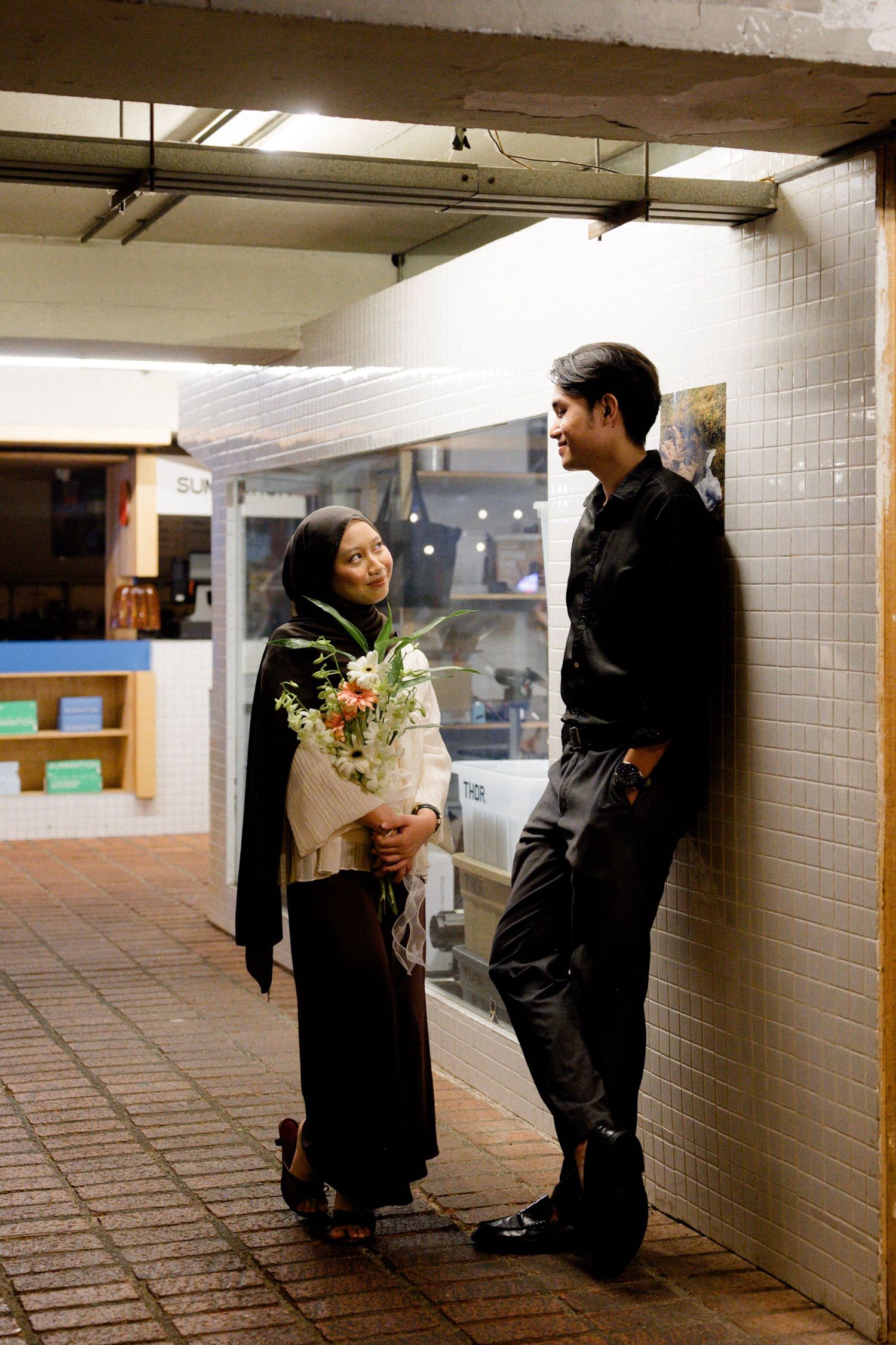 Muslim couple standing together in urban corridor, woman in hijab holding bouquet of white flowers.