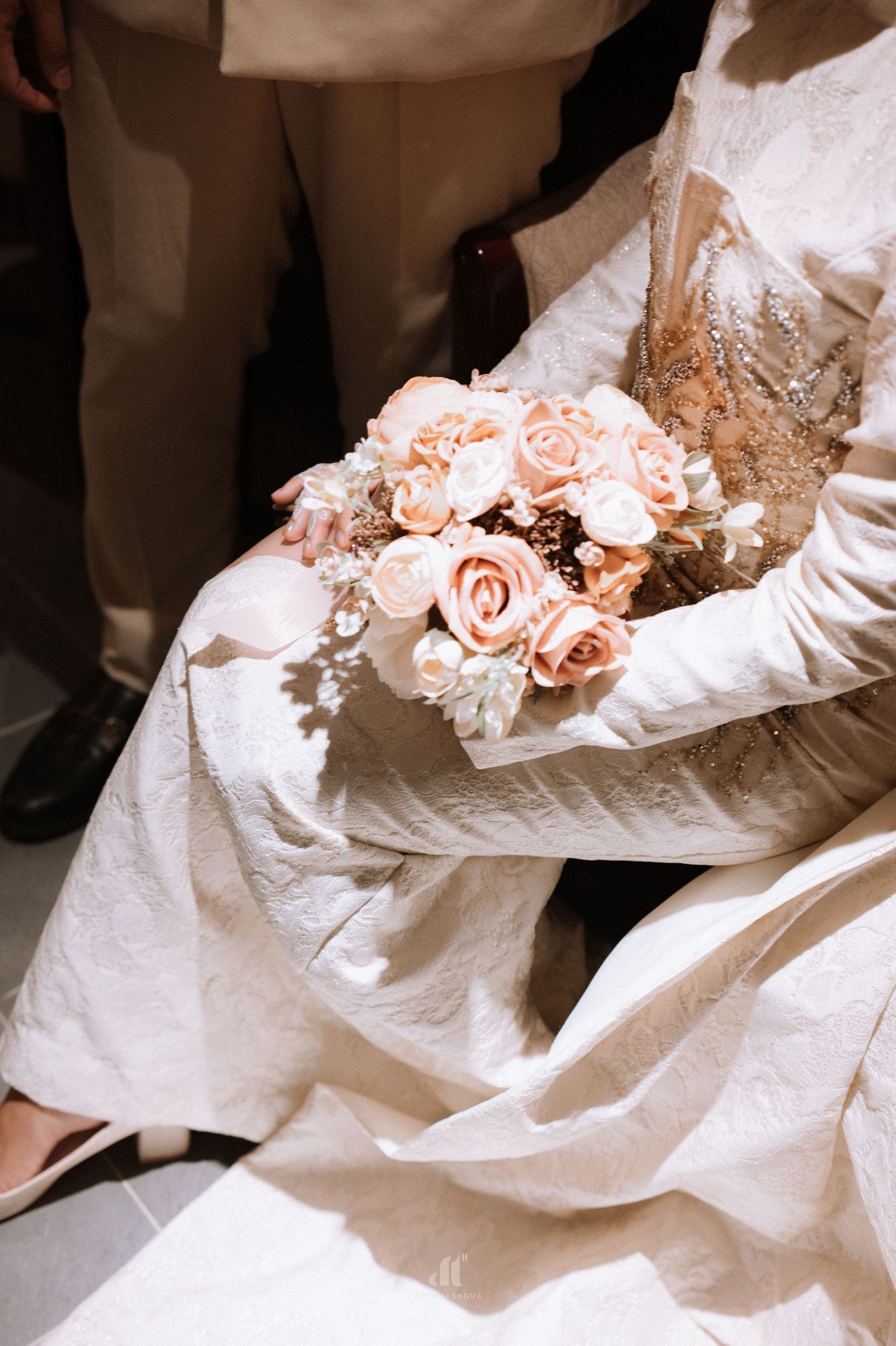 Bride in white lace gown holds a bouquet of pink and white roses on her lap.