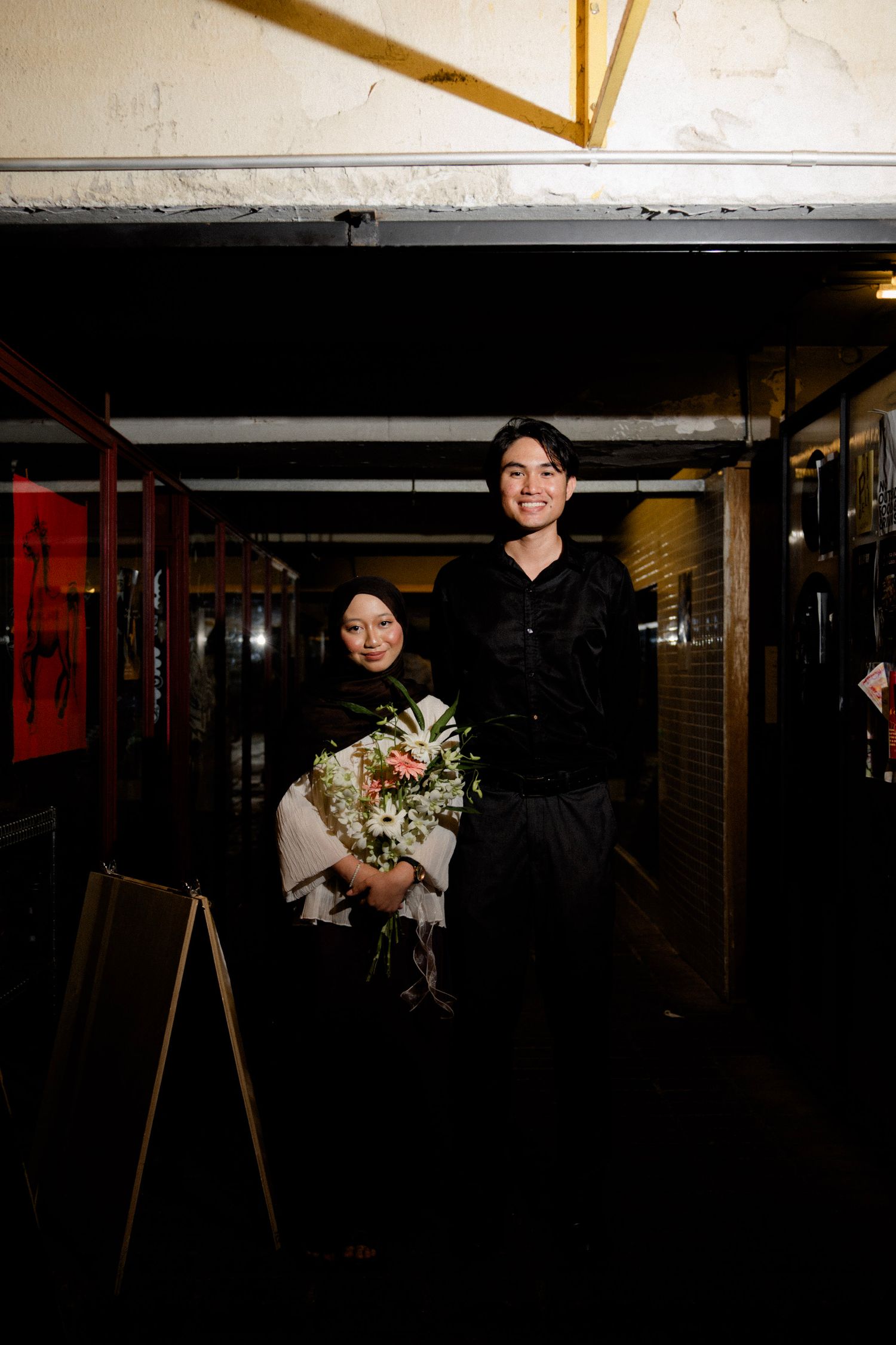 Smiling couple posing together at night with man in black and woman holding flower bouquet.