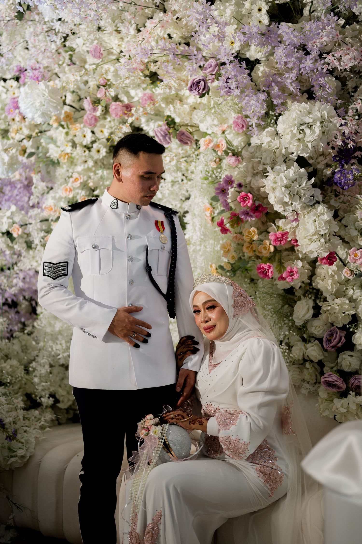 Groom in white military uniform stands beside seated bride in hijab amid floral backdrop.