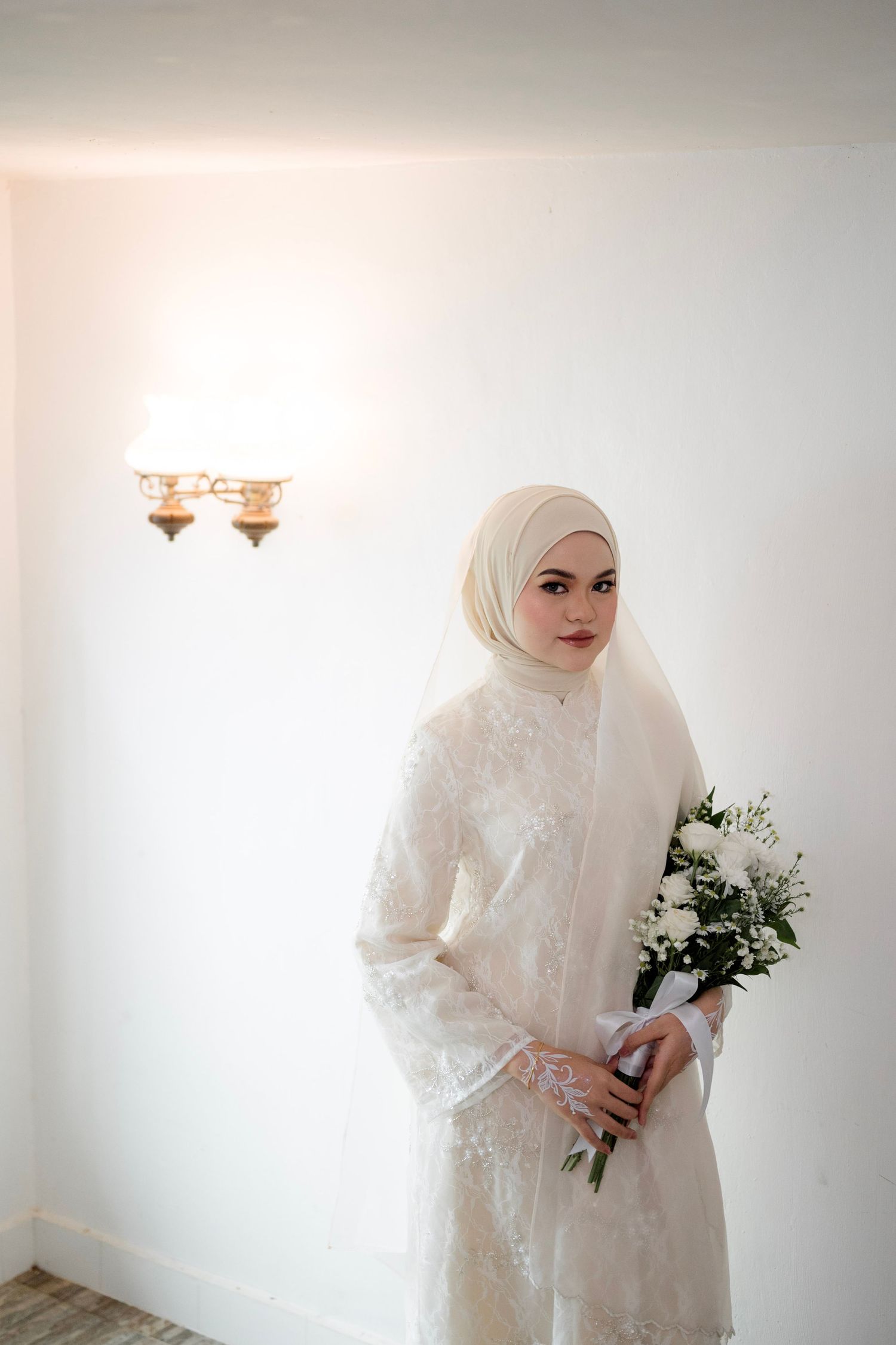 Muslim bride in white lace dress and hijab holding white rose bouquet against bright white wall.
