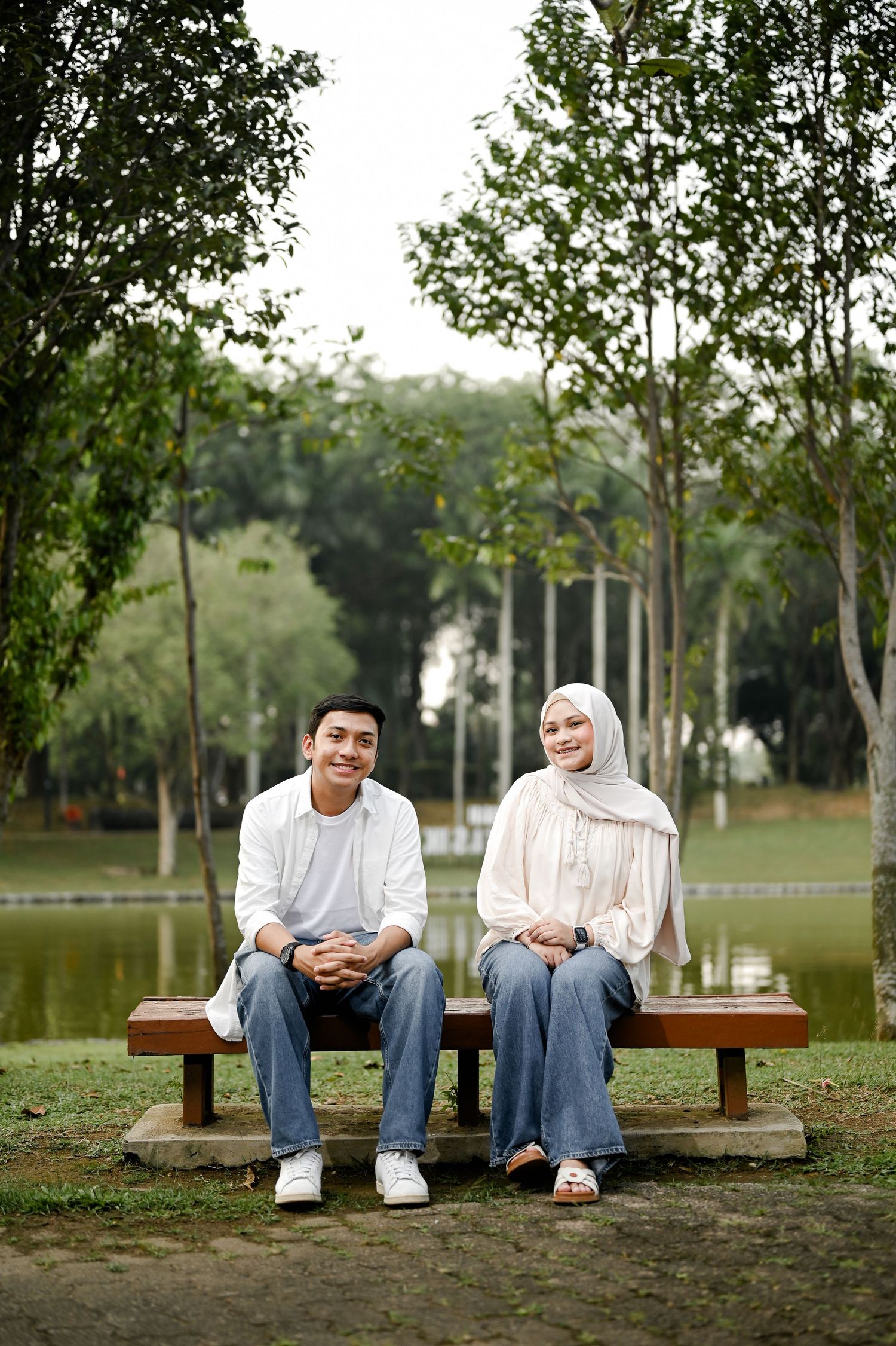 A young Muslim couple sitting on a park bench near a lake, smiling, wearing casual white tops and jeans.