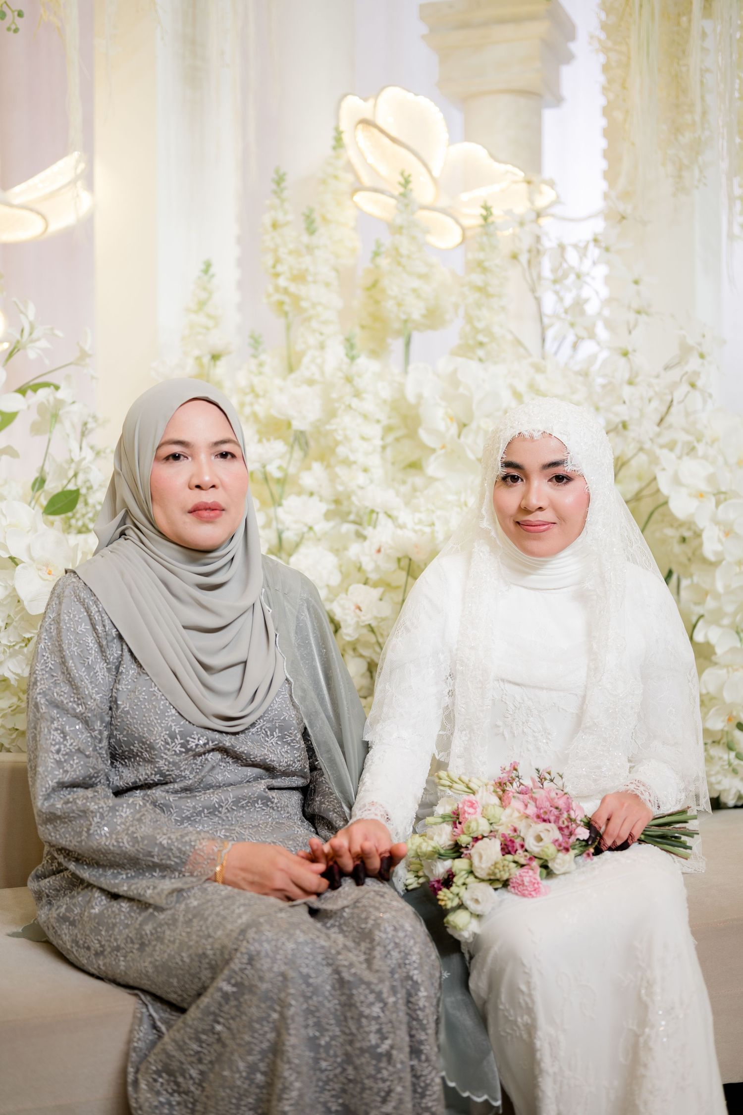 Muslim bride in white hijab holding pink bouquet seated with older woman in gray hijab at wedding.