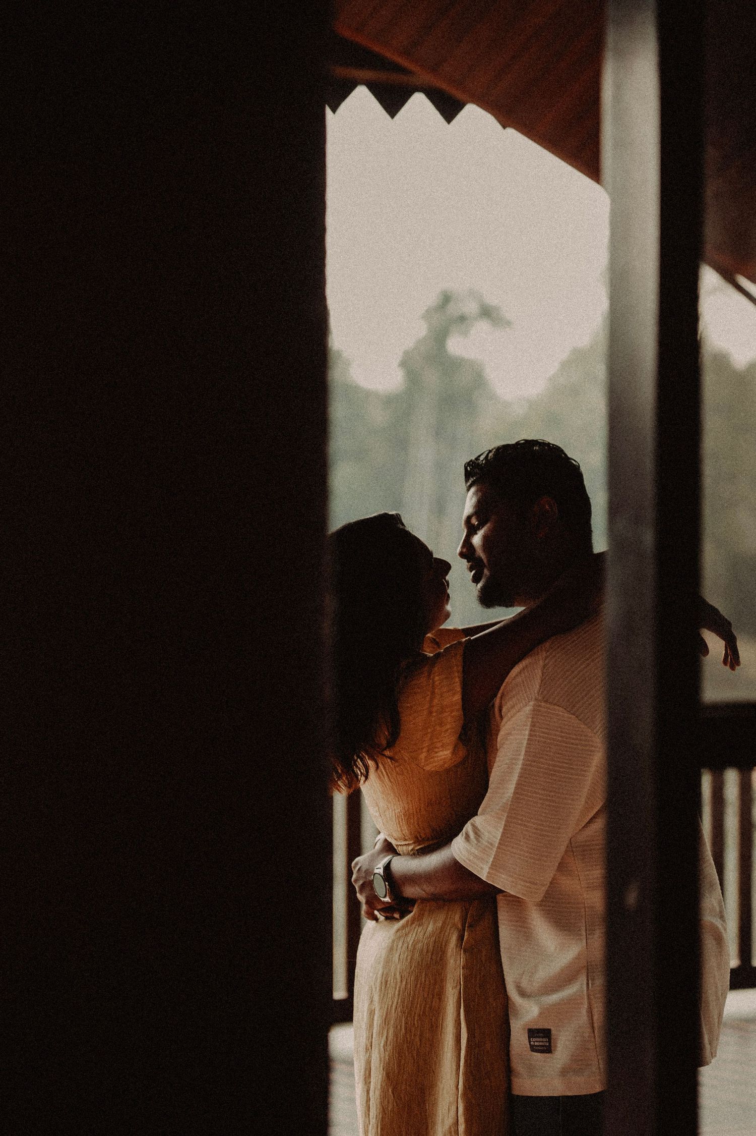 Romantic silhouette of couple embracing in doorway with lush green trees visible in background.