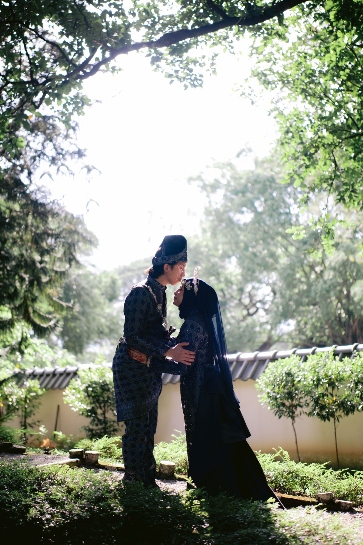 Couple in traditional attire sharing a romantic moment in a lush garden surrounded by green trees.