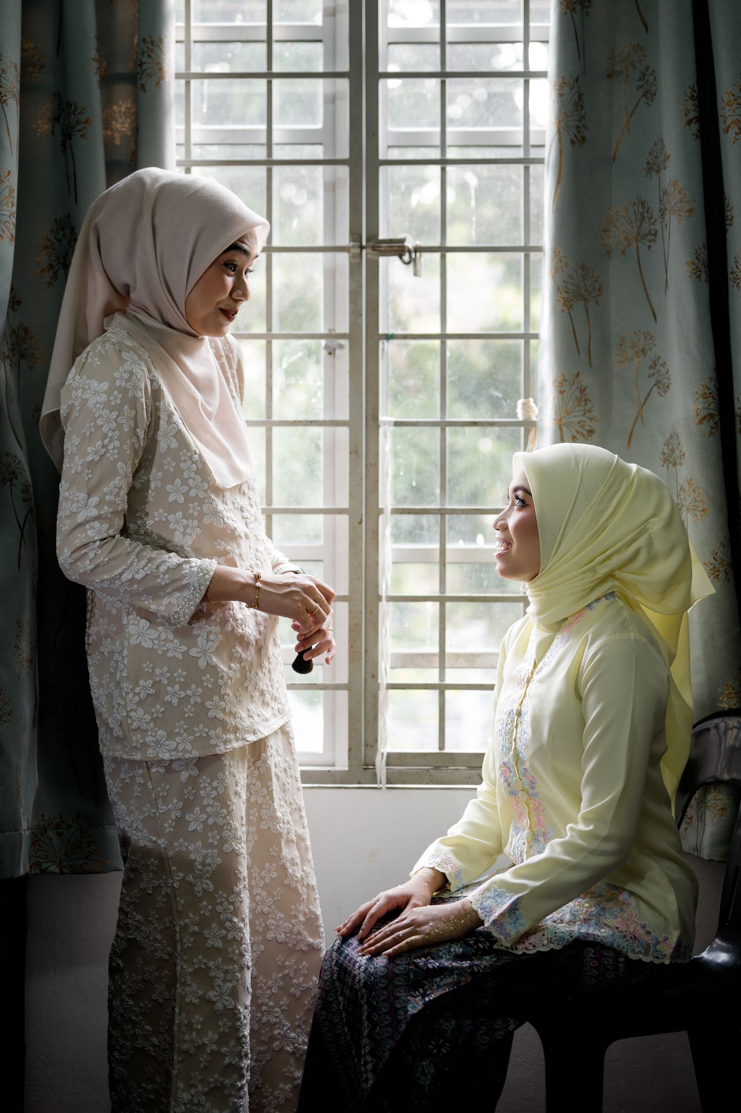 Two Muslim women in traditional Malay attire conversing by a latticed window with floral curtains.
