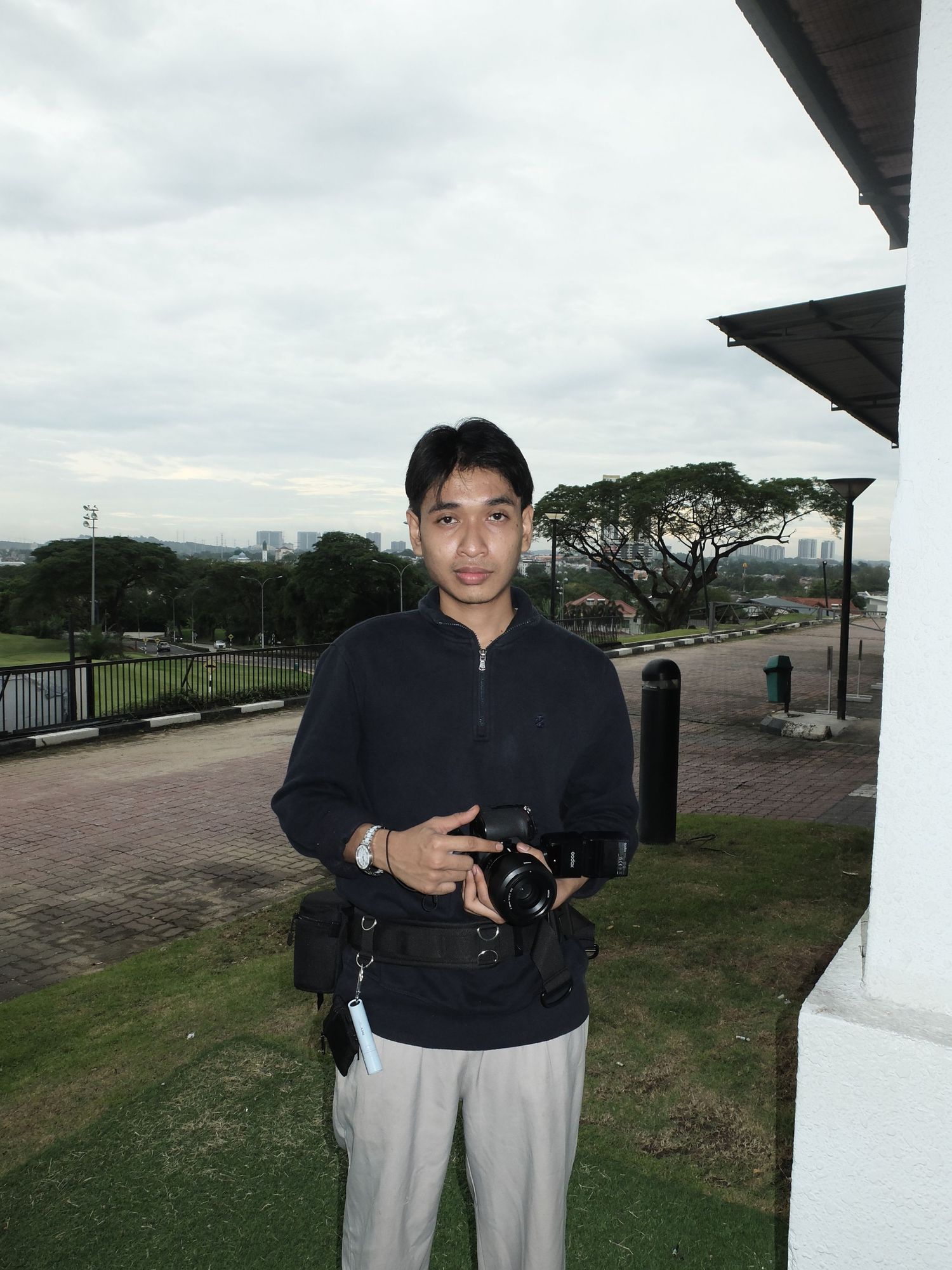 A young man in a dark shirt holds a professional camera outdoors near a building with a cityscape in the background.