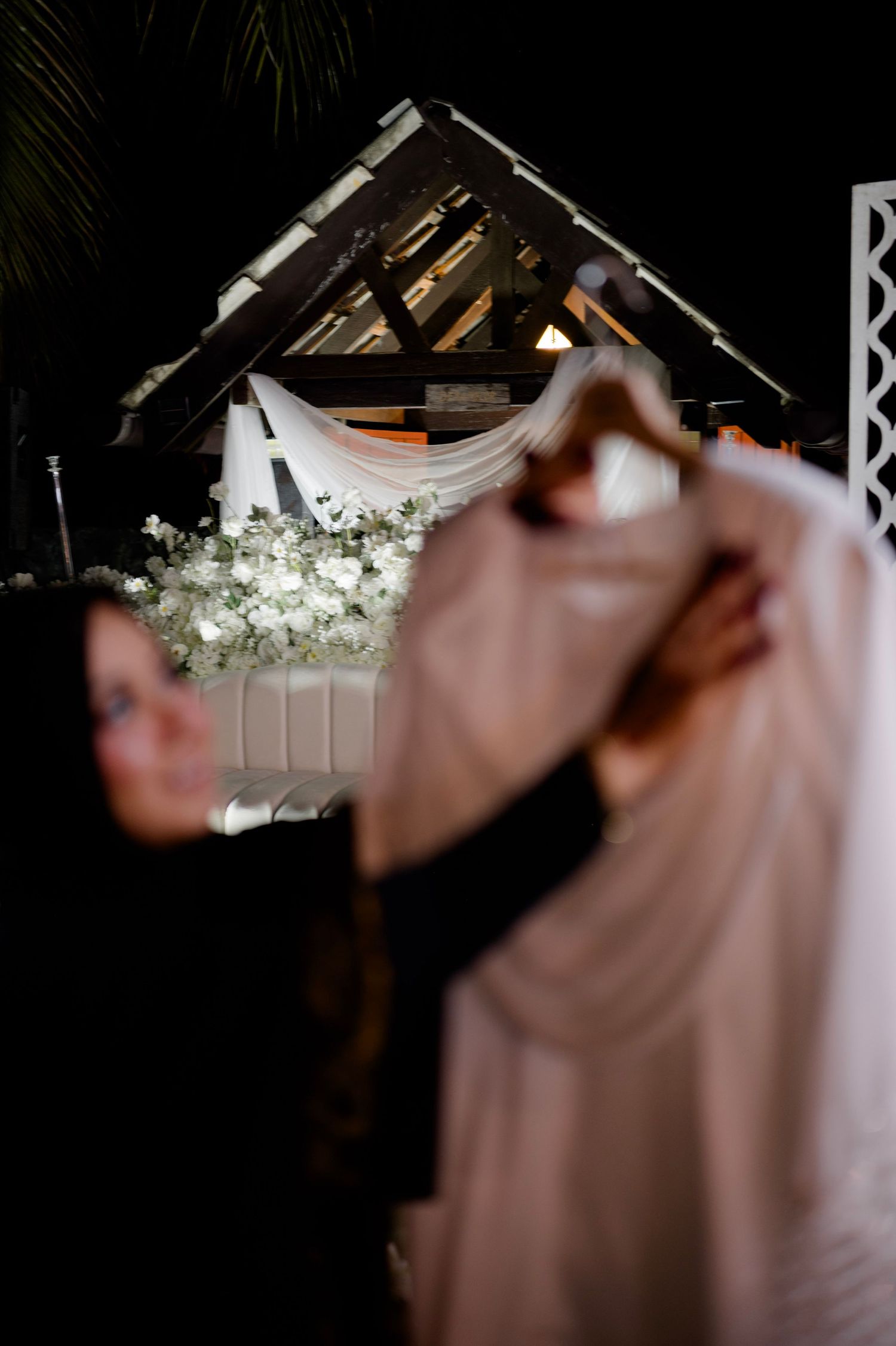 Blurred foreground of person holding dress hanger with decorated wedding arch in background.