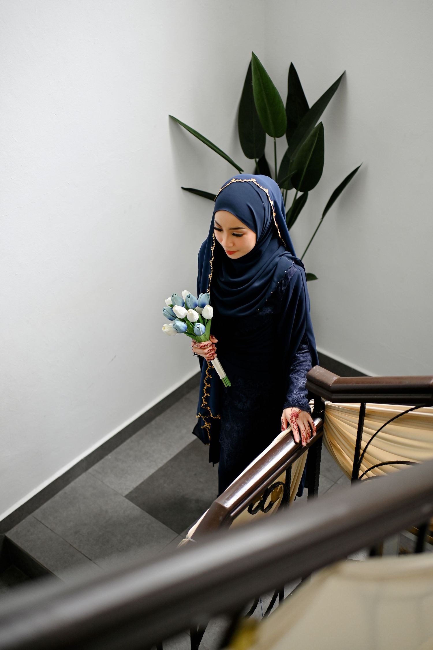 Muslim woman in navy hijab descending stairs holding blue and white flower bouquet in modern building.