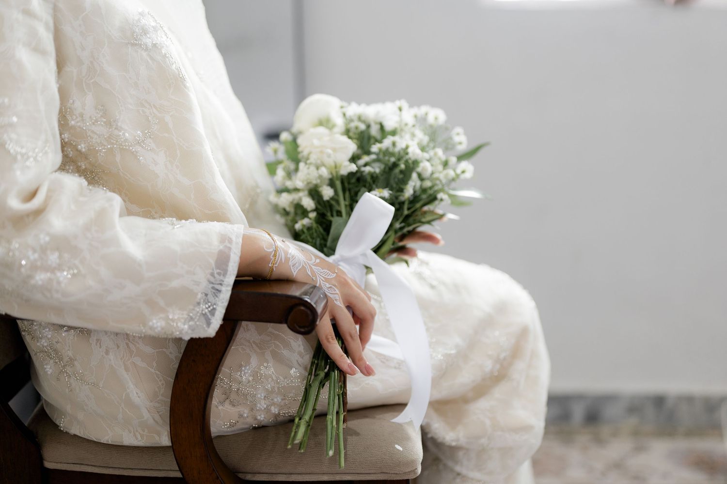 Bride in lace wedding dress sitting in wooden chair holding white floral bouquet tied with ribbon.