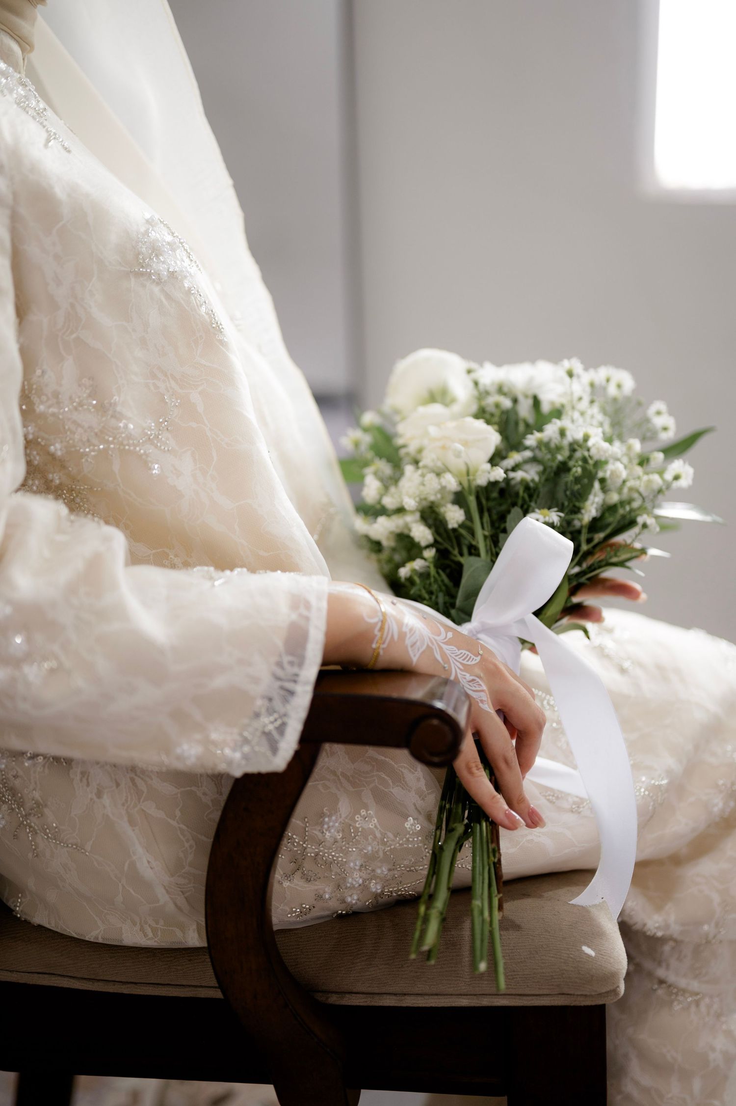 Close-up of bride in white lace dress holding elegant white bouquet with ribbon while seated in chair.