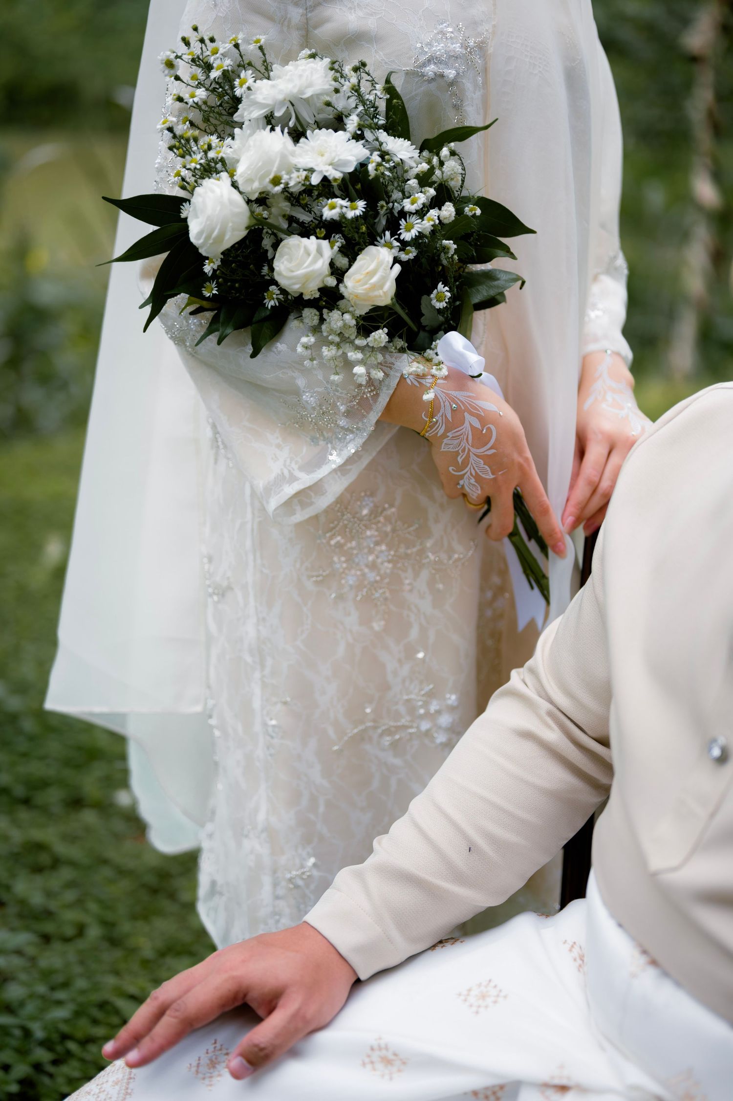 Bride in white lace dress holds a bouquet of white roses with henna-decorated hands.