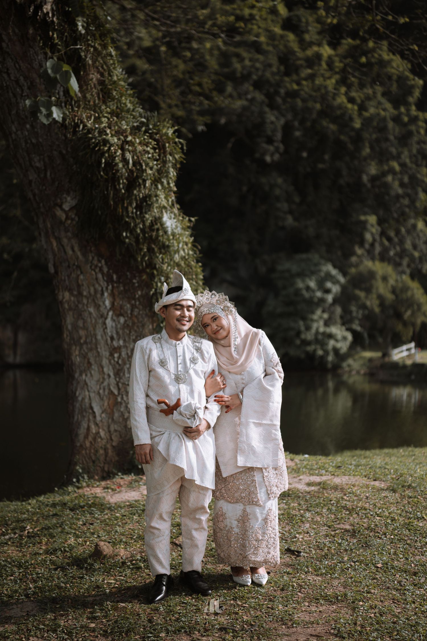 Muslim couple in white traditional attire posing outdoors near a tree and lake.