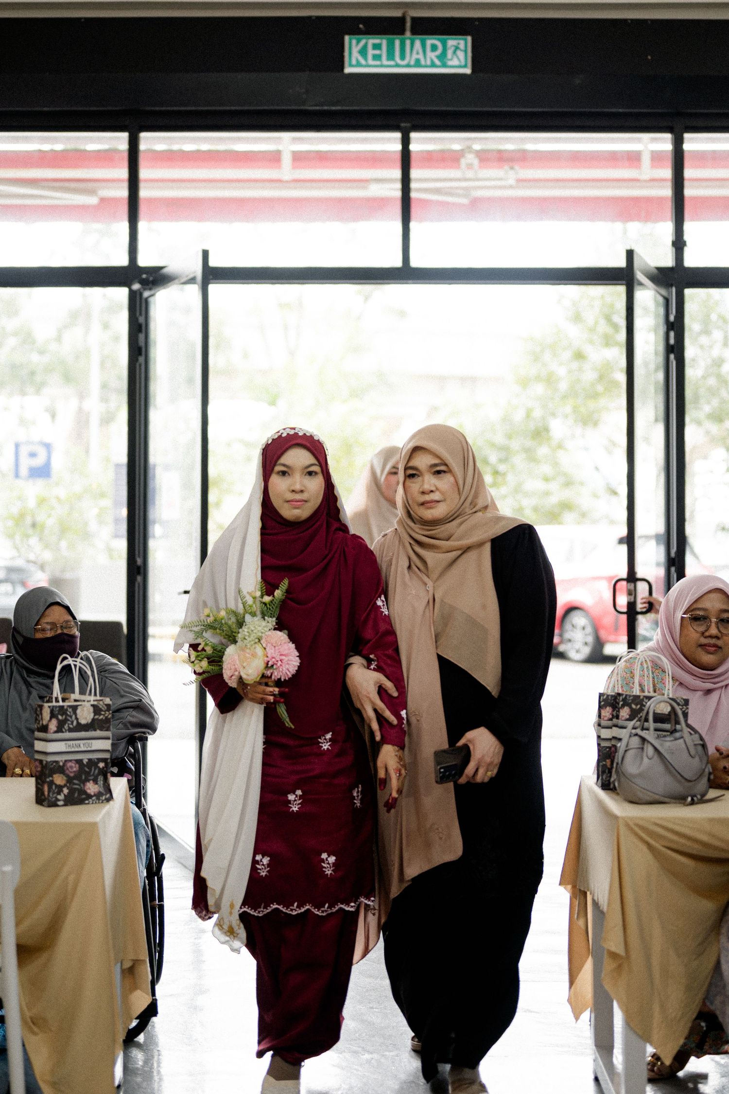 Two Muslim women in hijabs walk through an exit door, one holding a bouquet of pink flowers at a formal event.