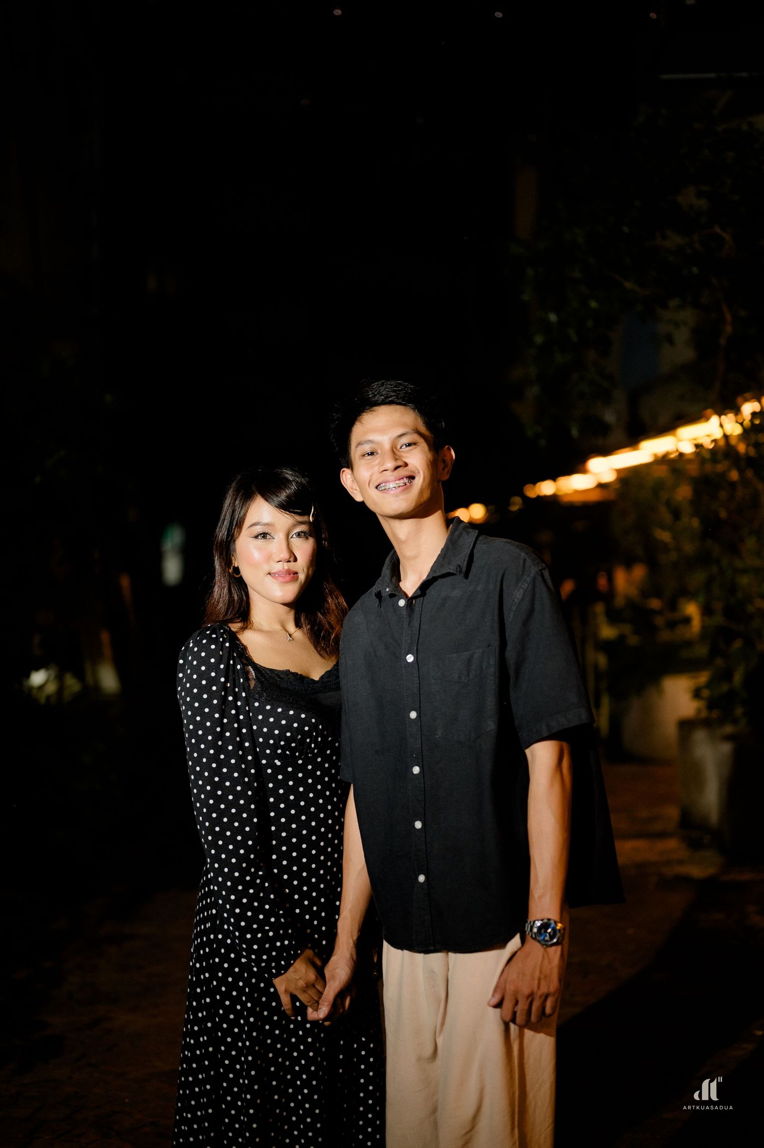 Couple holding hands and smiling together outdoors at night with warm string lights behind.