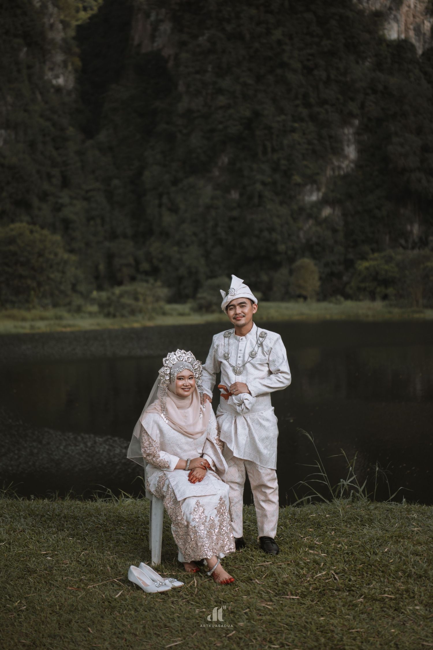 Malaysian couple in traditional Malay wedding attire posing outdoors near a lake with lush greenery and limestone cliffs.