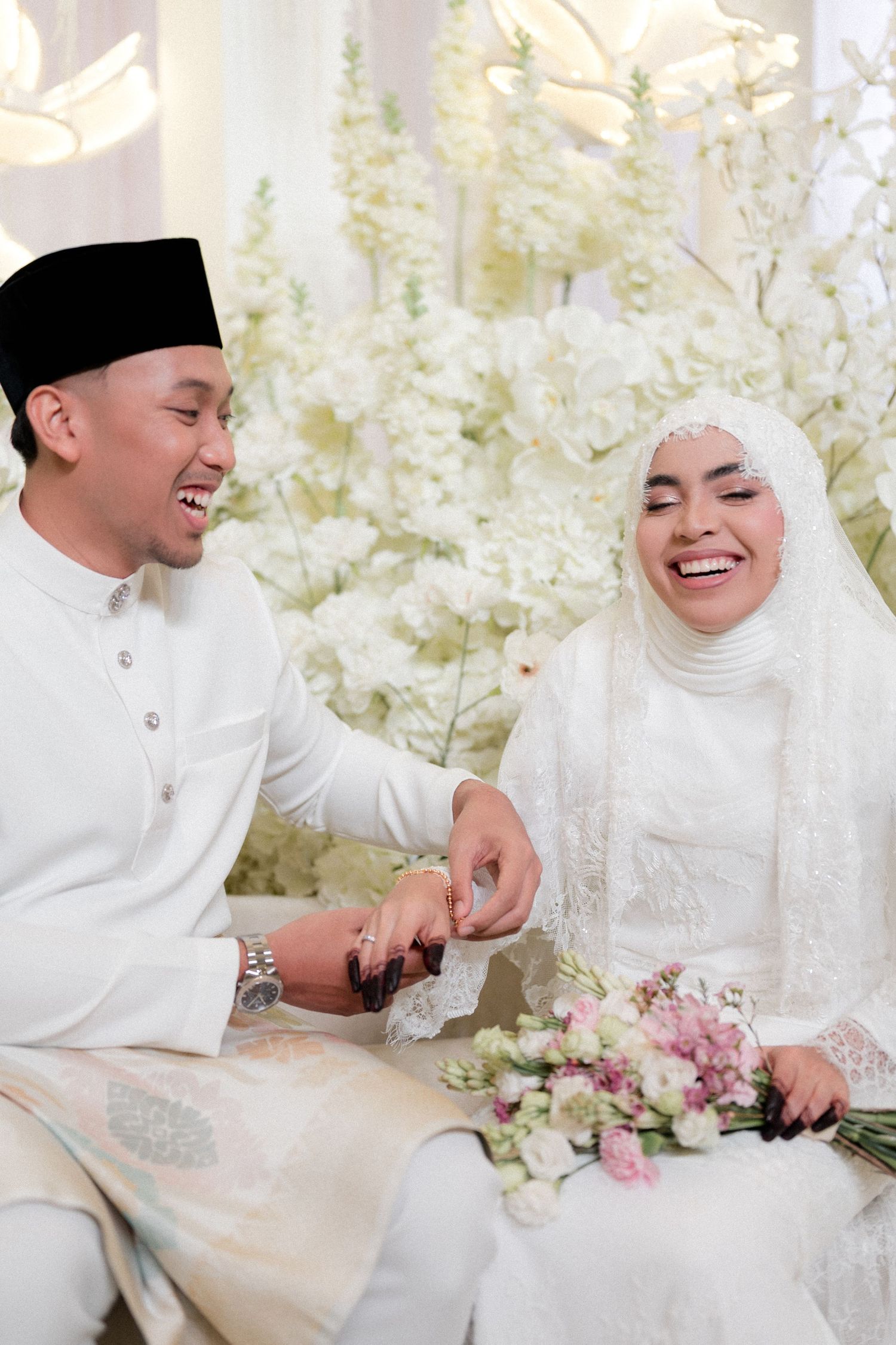 Malaysian Muslim couple in white traditional attire laughing together at wedding reception with floral backdrop.