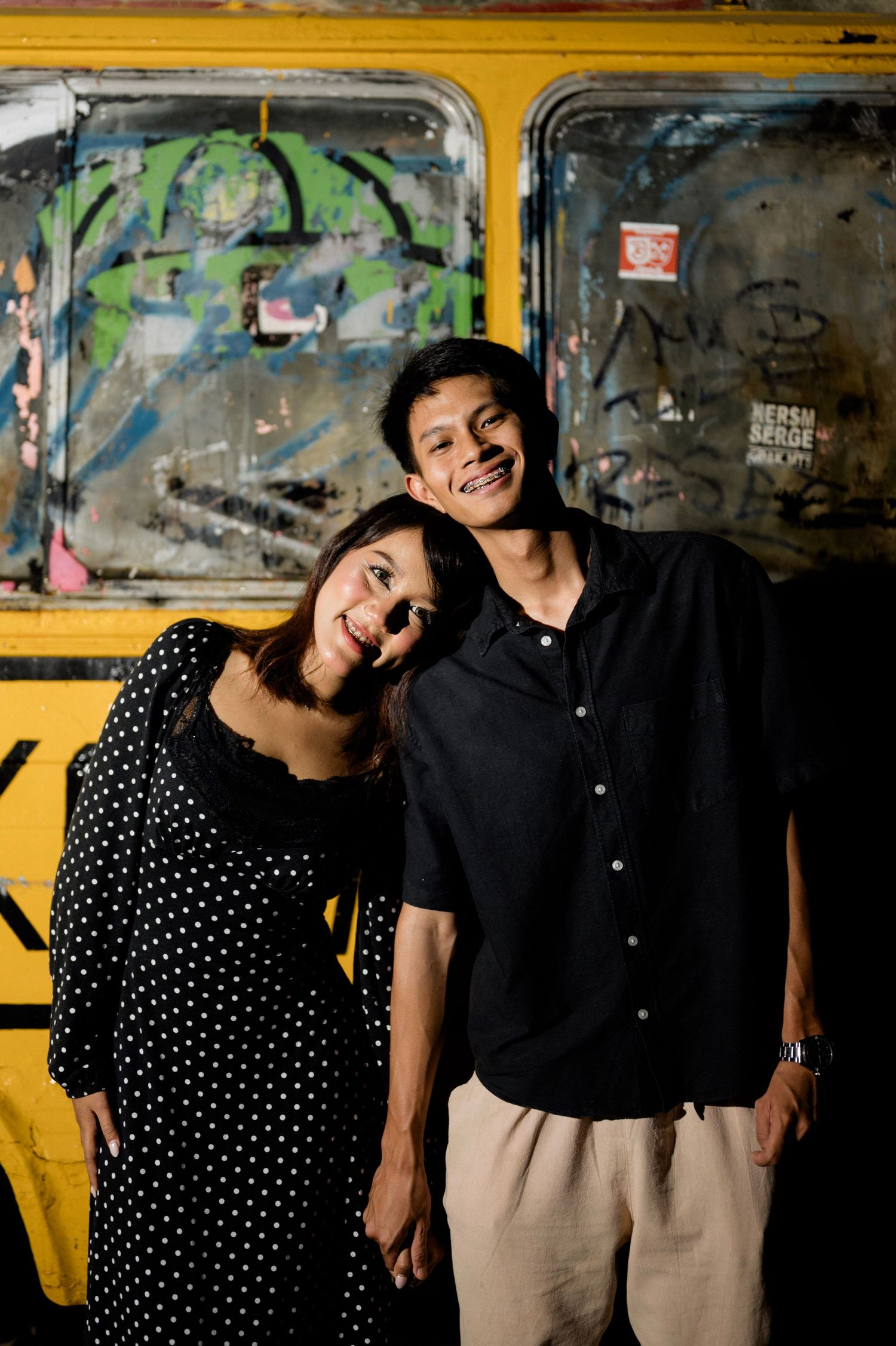 Smiling Asian couple posing together in front of colorful graffiti-covered yellow bus at night.