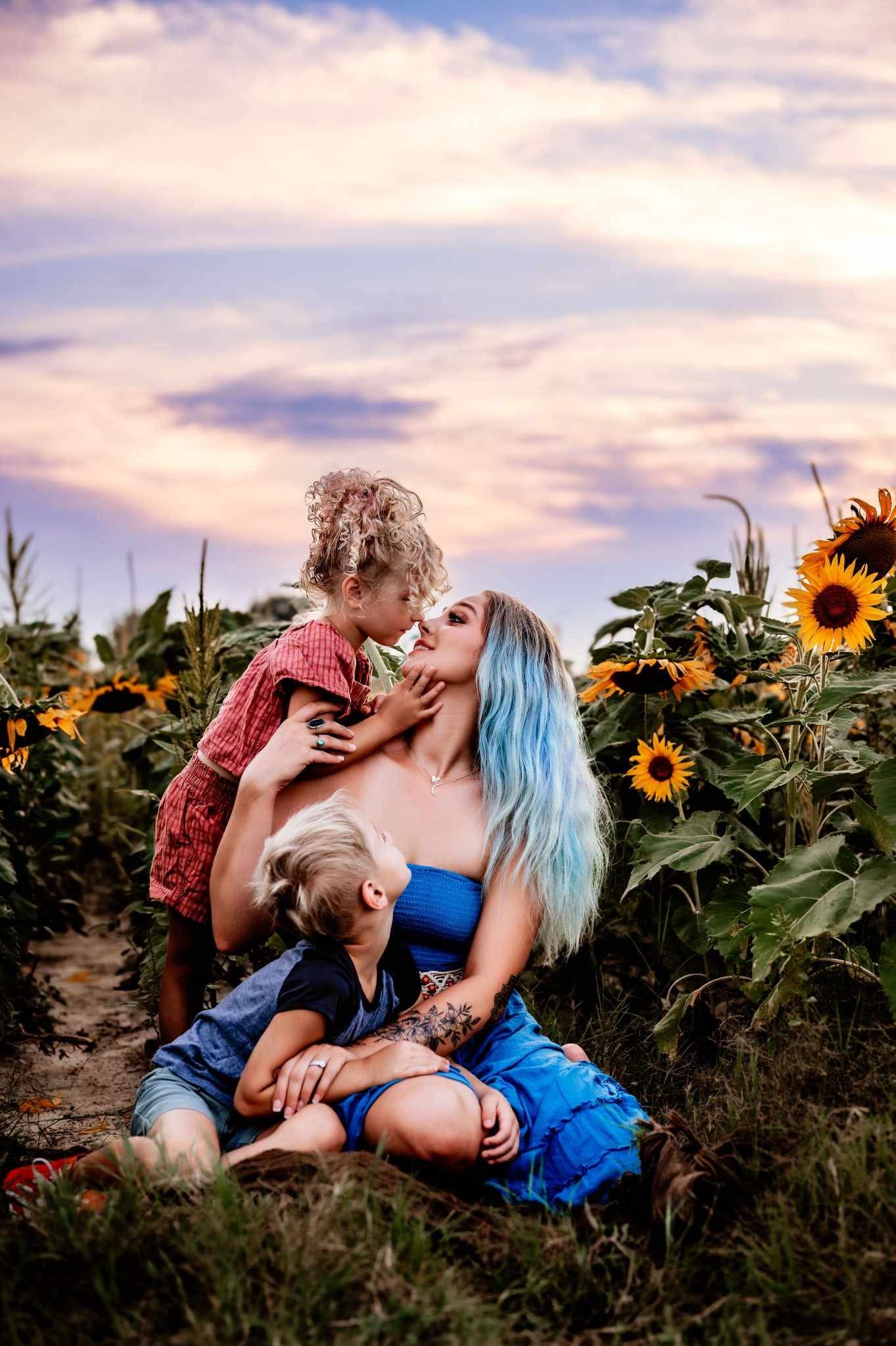 Mom snuggles her two little ones in a sunflower field beneath a vibrant cotton candy sky with colorful, dreamy, slightly moody tones.