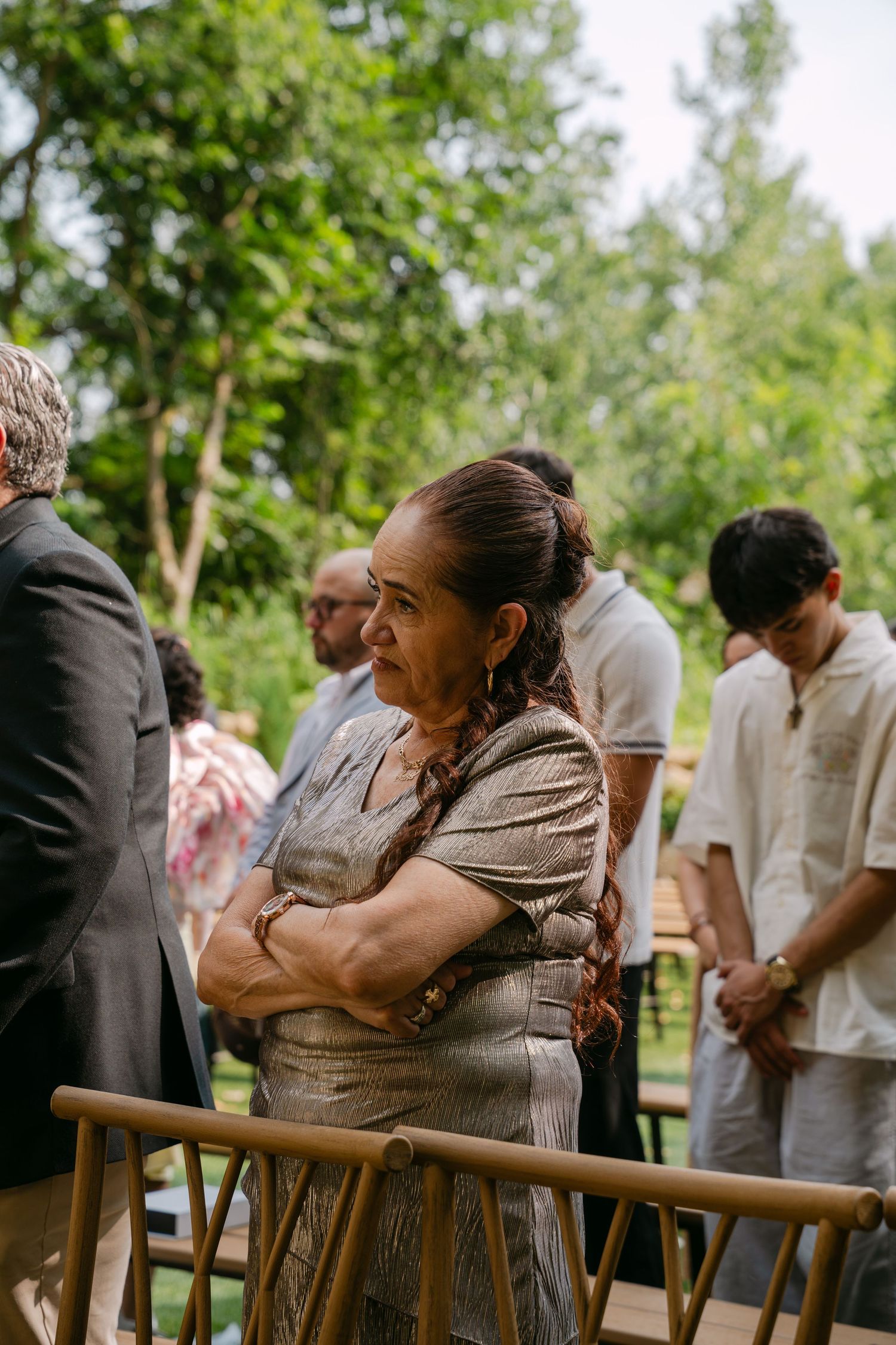 Elderly woman in silver dress with arms crossed watches emotional outdoor wedding ceremony.