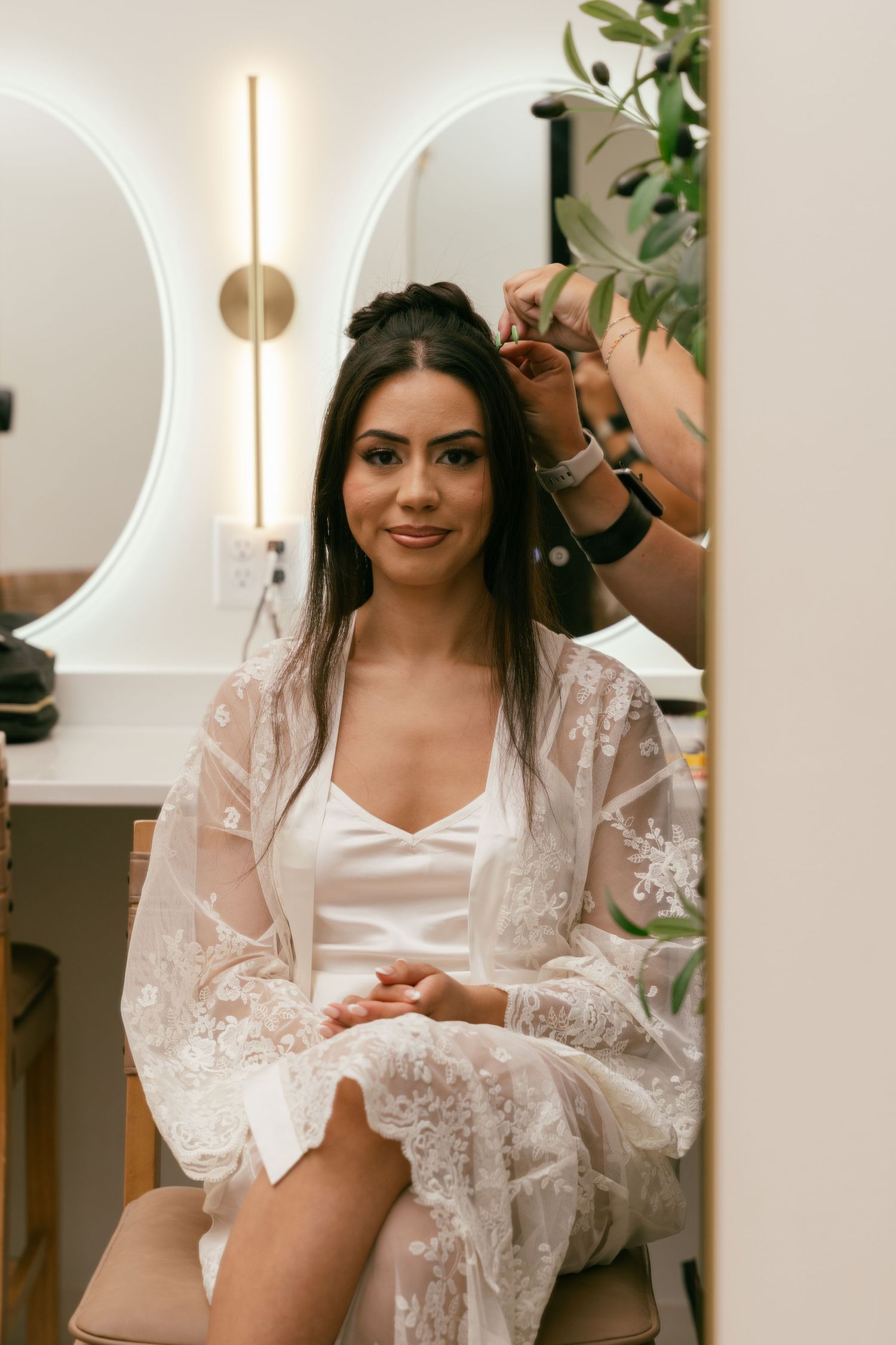 Bride smiling while getting hair styled in bridal suite wearing white lace robe before wedding ceremony.