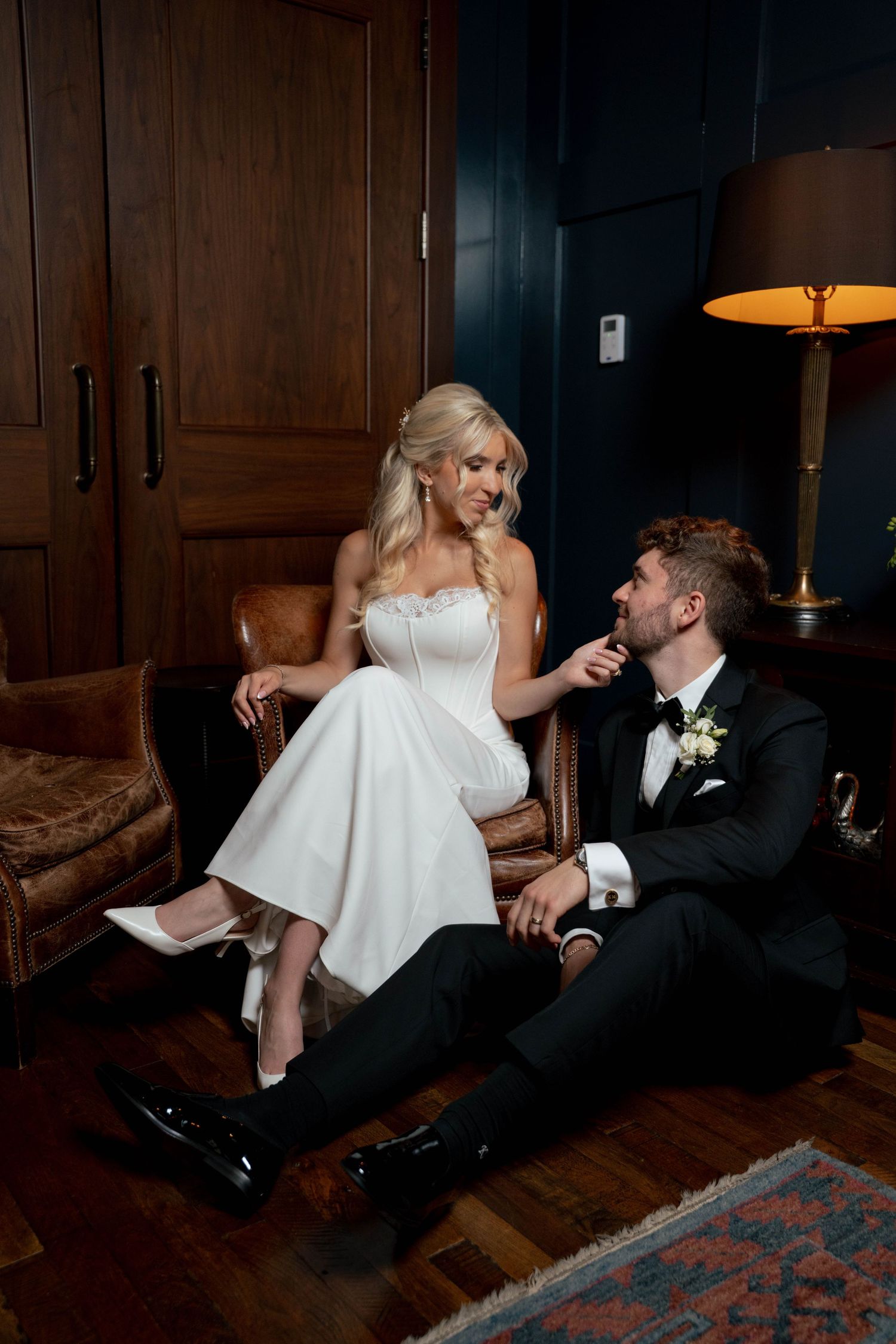 Bride in white strapless gown sits on leather chair while groom in black tuxedo sits on floor gazing at her lovingly.