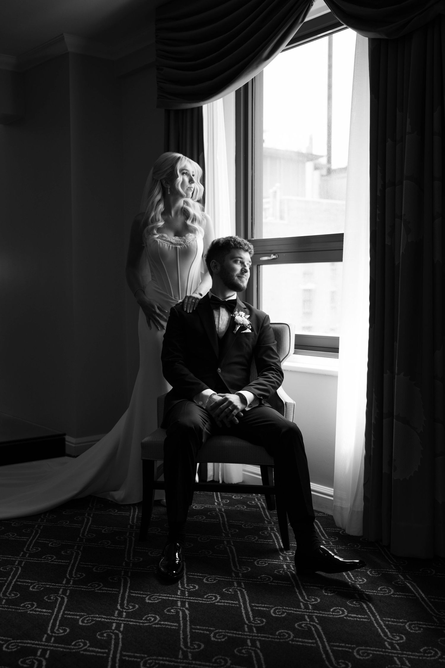 Black and white photo of bride and groom posing by a window in a luxurious hotel room.