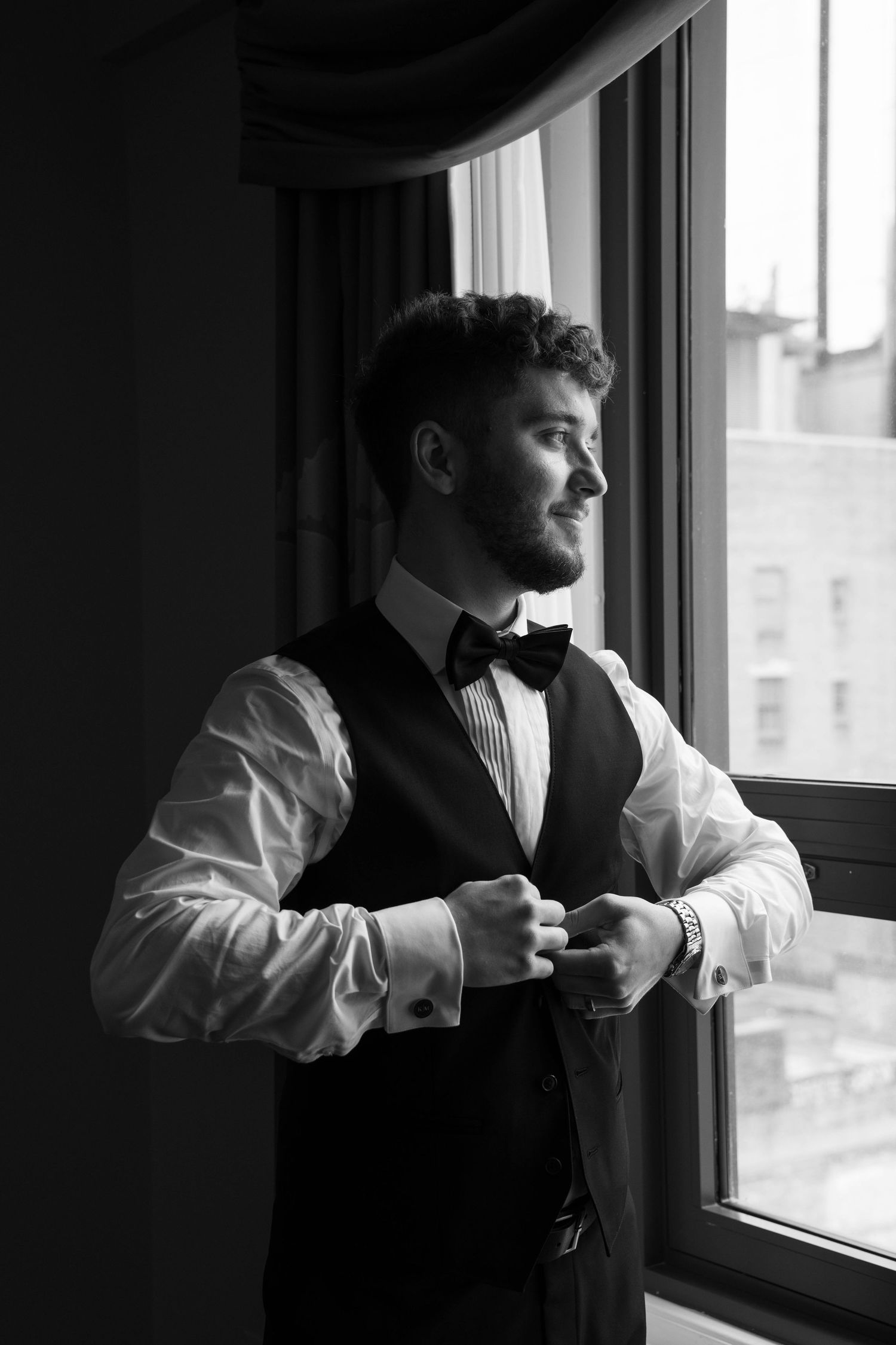 Dapper groom in black vest and bow tie gazing out hotel window before wedding ceremony.