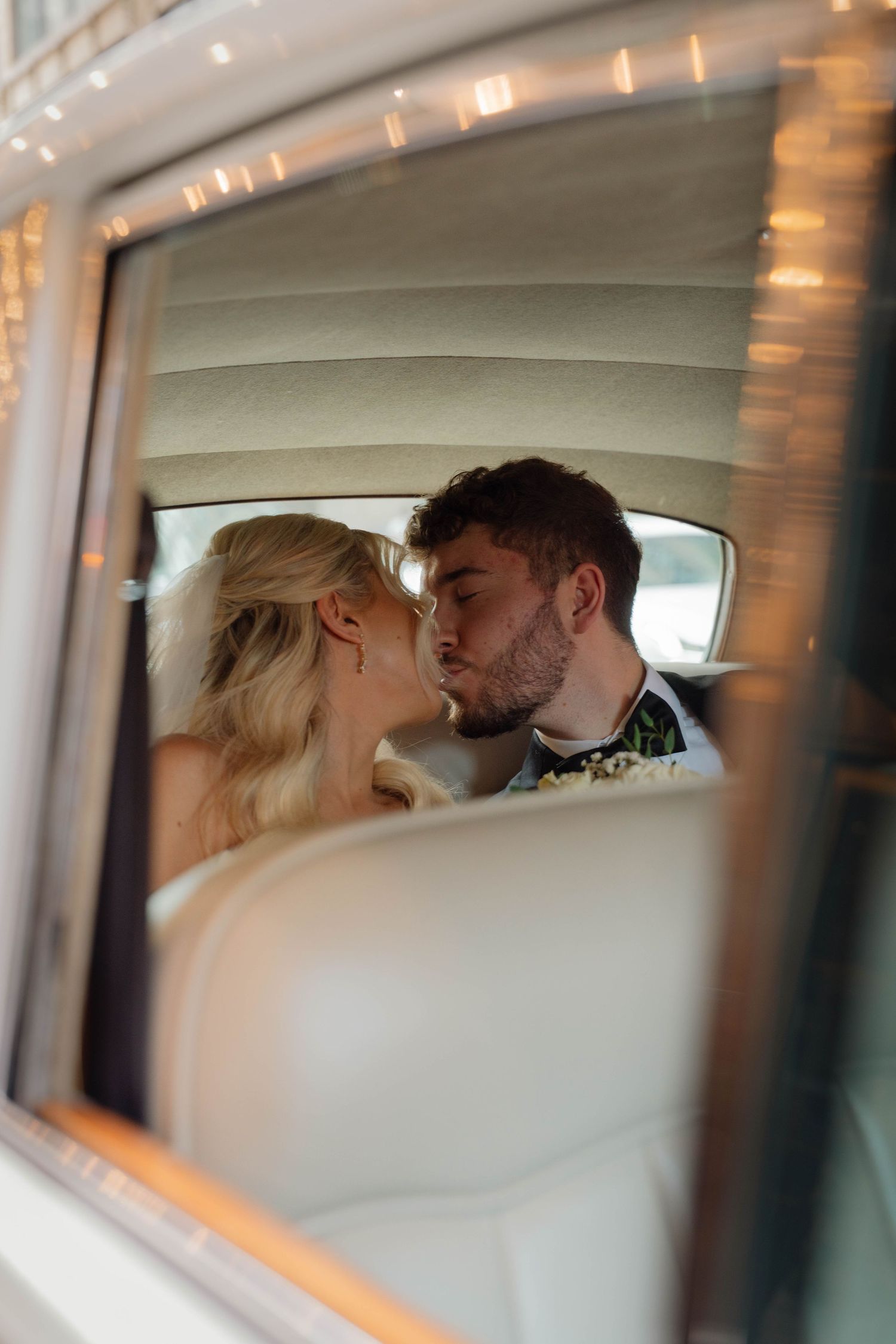 Bride and groom sharing romantic kiss in vintage car backseat with fairy lights.