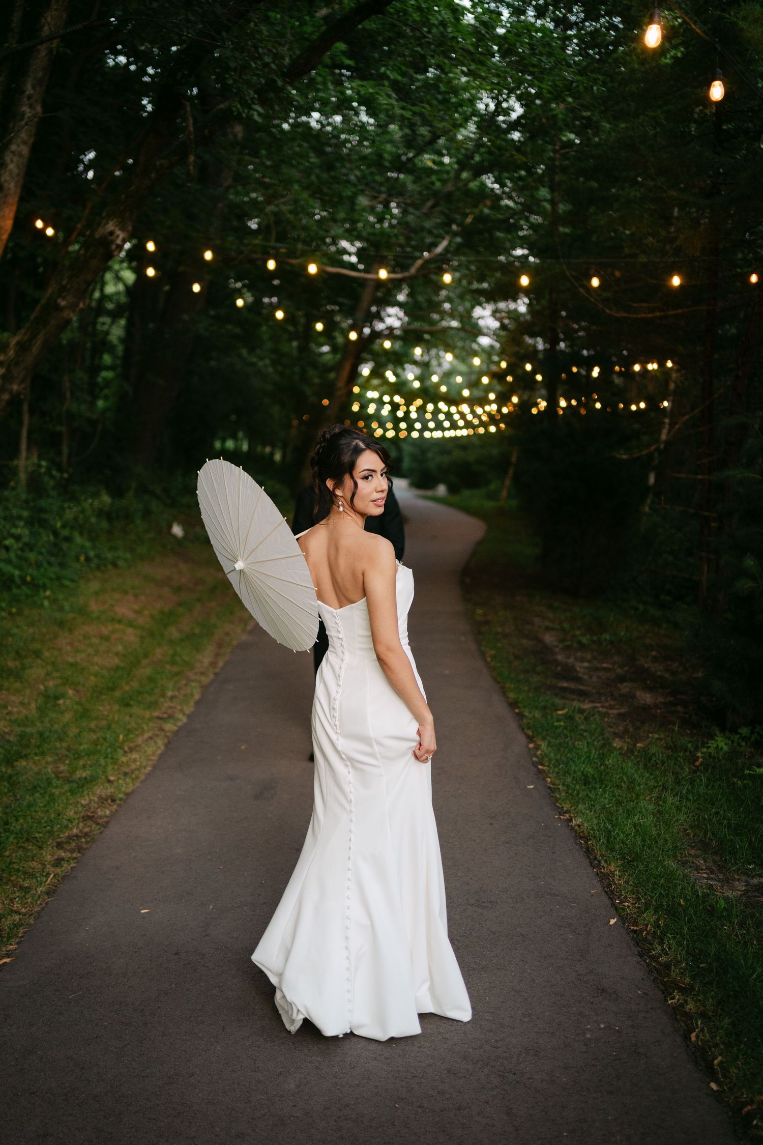 Bride in white wedding gown holds parasol while walking down tree-lined path with string lights.