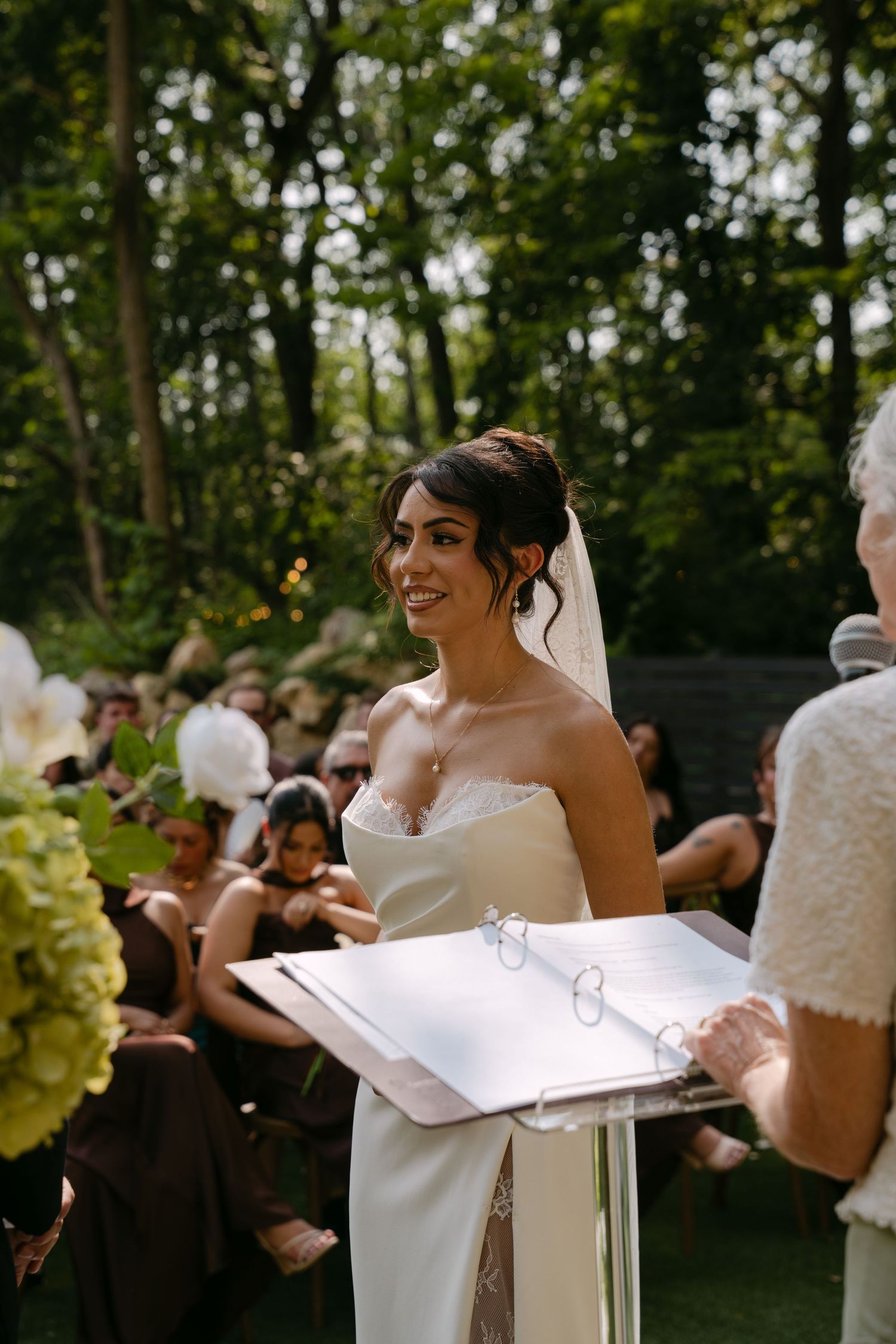 Smiling bride in white strapless gown reads vows at outdoor wedding ceremony with guests seated behind.