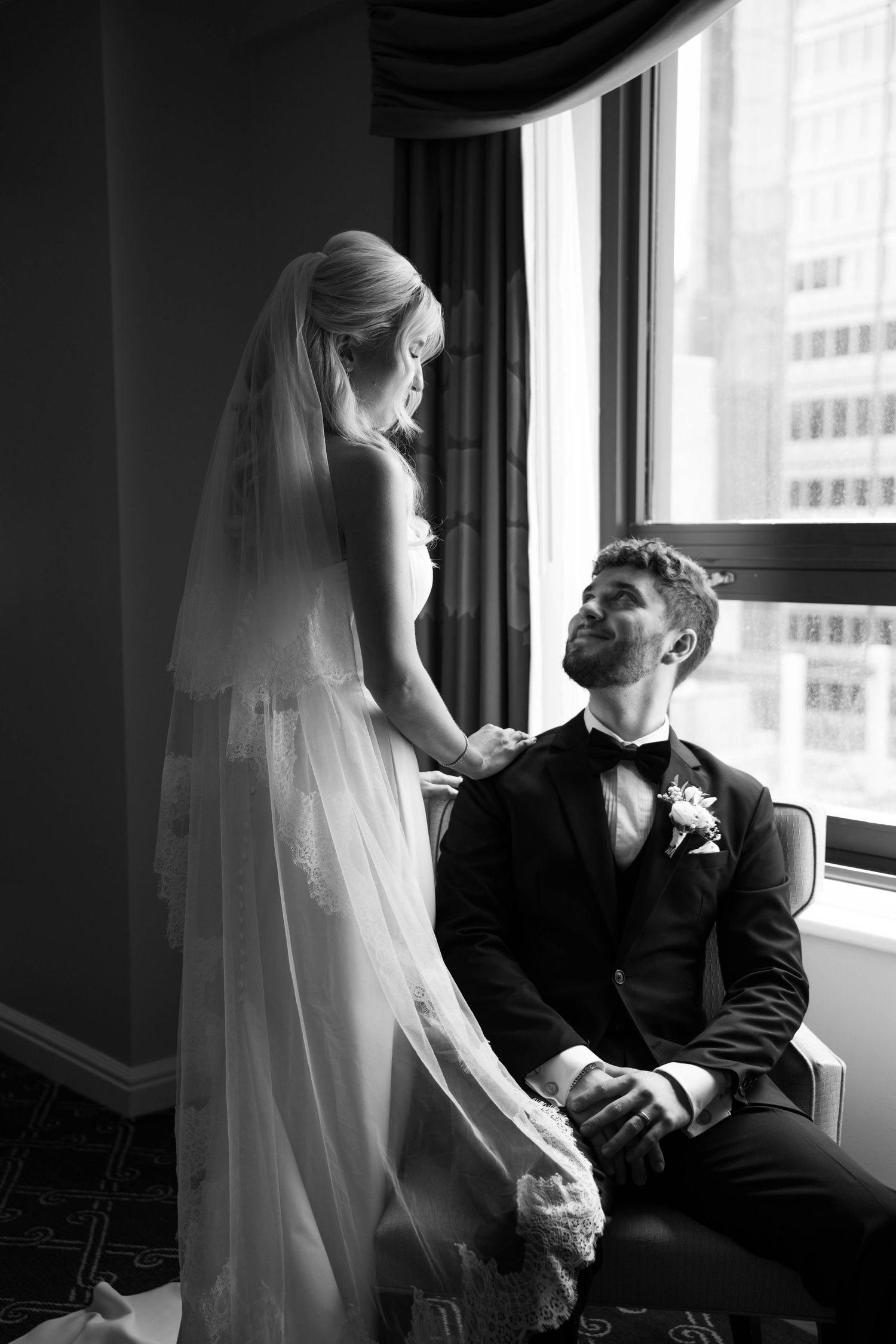 Groom gazes up at bride in lace veil by a sunlit window in black and white.