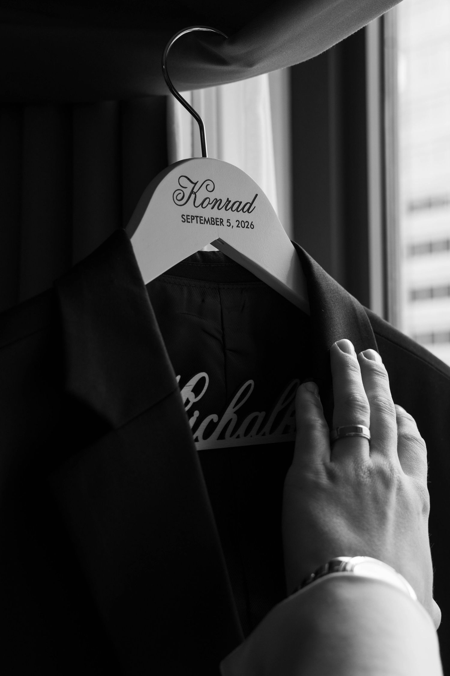 Black and white photo of a personalized wedding hanger reading Konrad September 5, 2026 with a suit jacket.