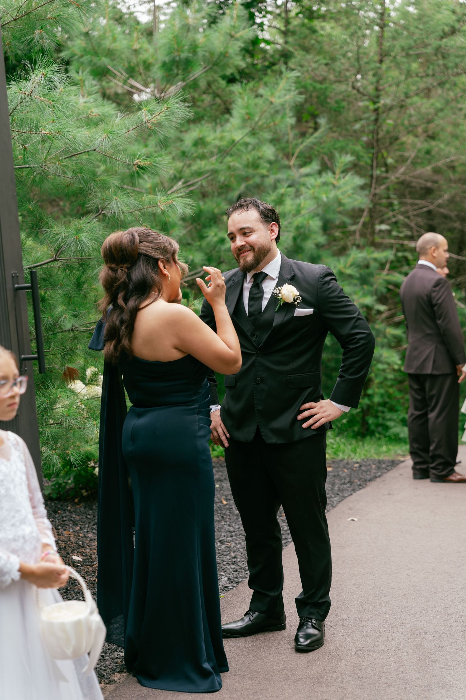 Groomsman in black suit smiling while chatting with bridesmaid in teal gown at outdoor wedding.