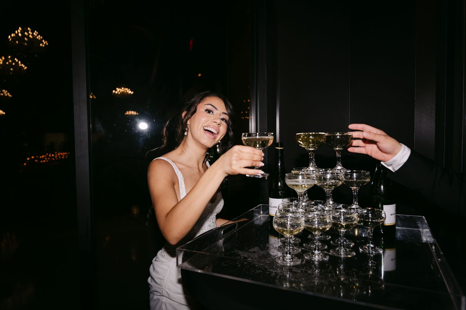 Smiling bride in white mini dress pours champagne tower while guest reaches for a glass.