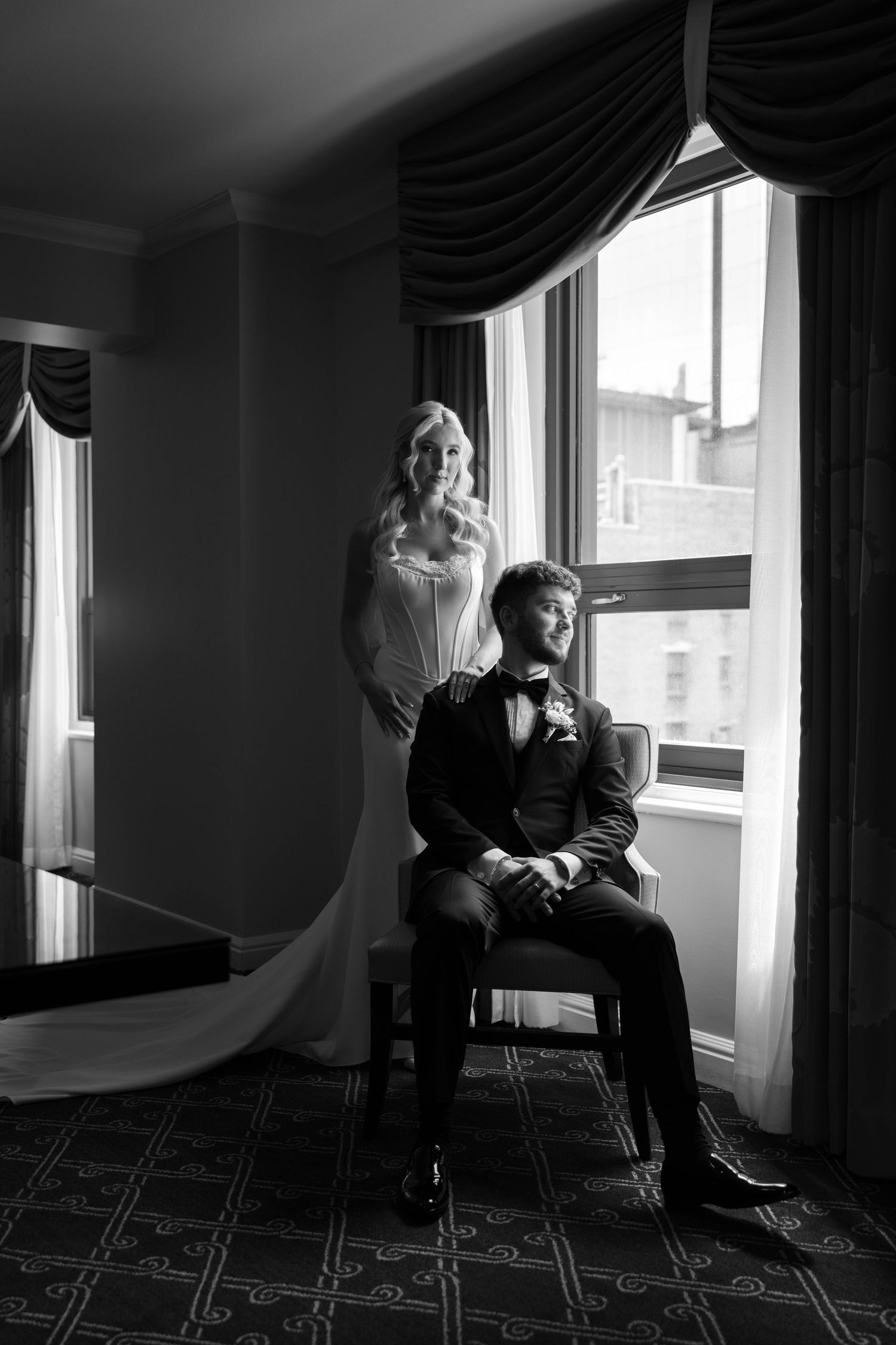 Black and white wedding photo of bride standing behind seated groom near elegant hotel window with draped curtains.