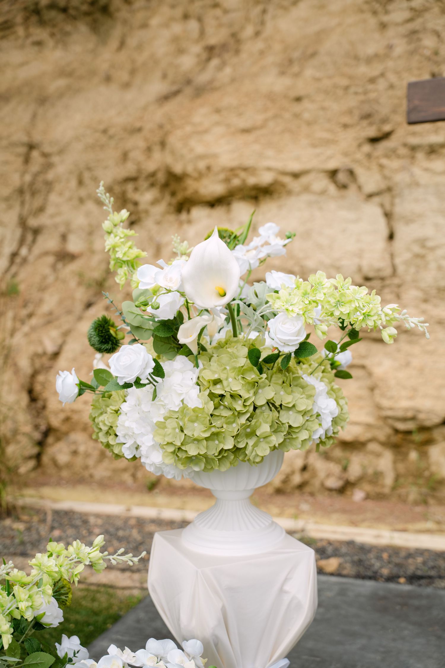 White urn vase filled with hydrangeas, roses, and calla lilies displayed against a stone wall.