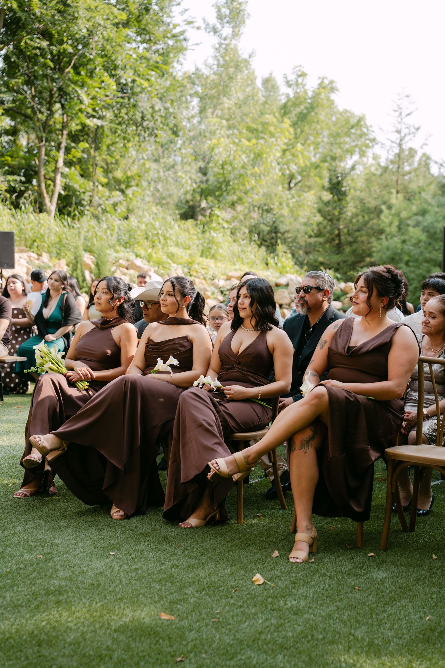 Four bridesmaids in brown dresses seated together at outdoor garden wedding ceremony.